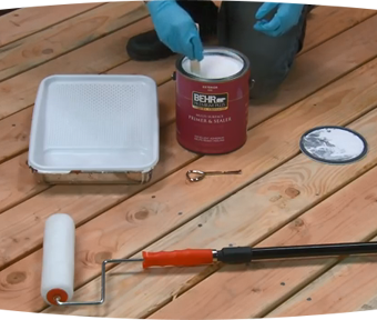 A person kneeling down with paint brush in hand and paint roller and paint can aside them