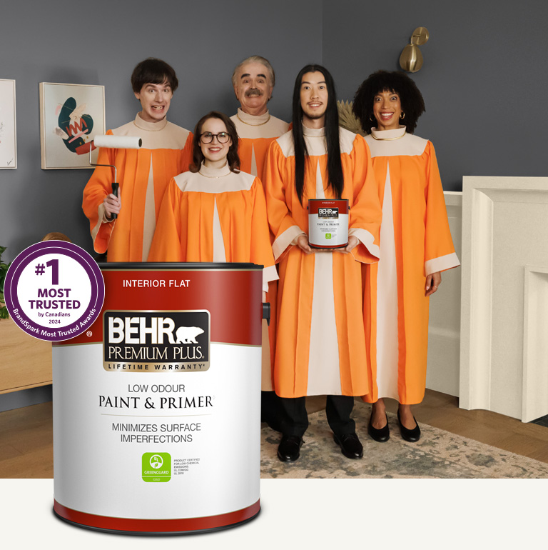 Mobile version of a choir standing in a painted living room and holding a can of Behr Premium Plus Interior Flat Paint