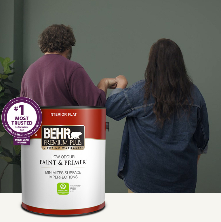Mobile version of a choir standing in a painted living room and holding a can of Behr Premium Plus Interior Flat Paint