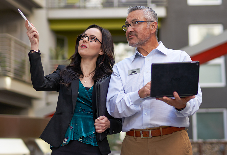A small image of a Property Manager showing a BEHR PRO Rep a multi-family complex. The property manager is pointing at something while the BEHR PRO Rep is looking at what she is pointing and taking notes on a laptop.