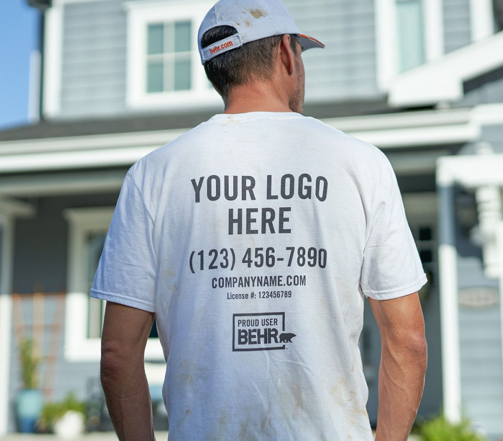 Desktop view of  Pro facing a house. The back of his shirt is on focus with the words in-printed Your Logo here - Fake Telephone # - companyname.com and a logo of PROUD USER BEHR.