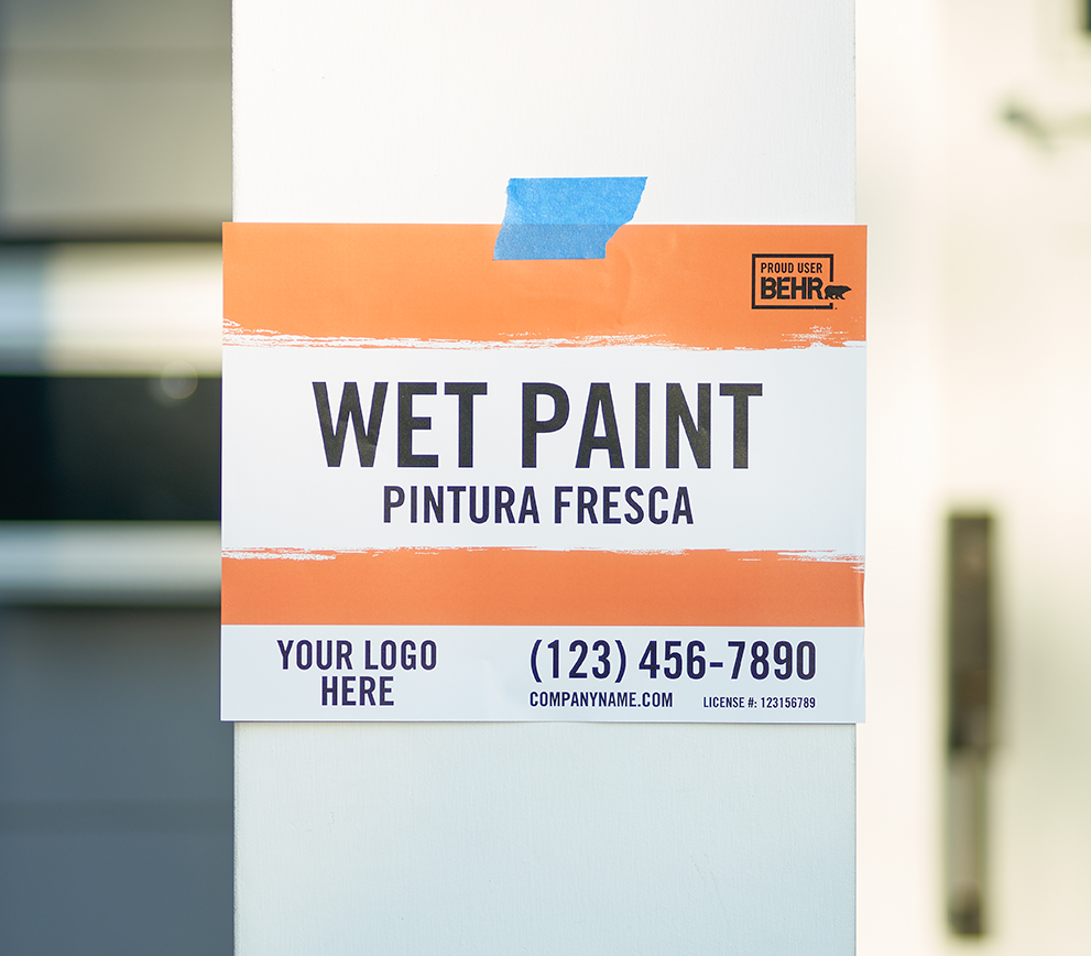 A close up view of a sign that is stuck to a column of a house with the words printed WET PAINT - PINTURA FRESCA - your logo here- (123) 456-7890 - companyname.com - PROUD USER BEHR