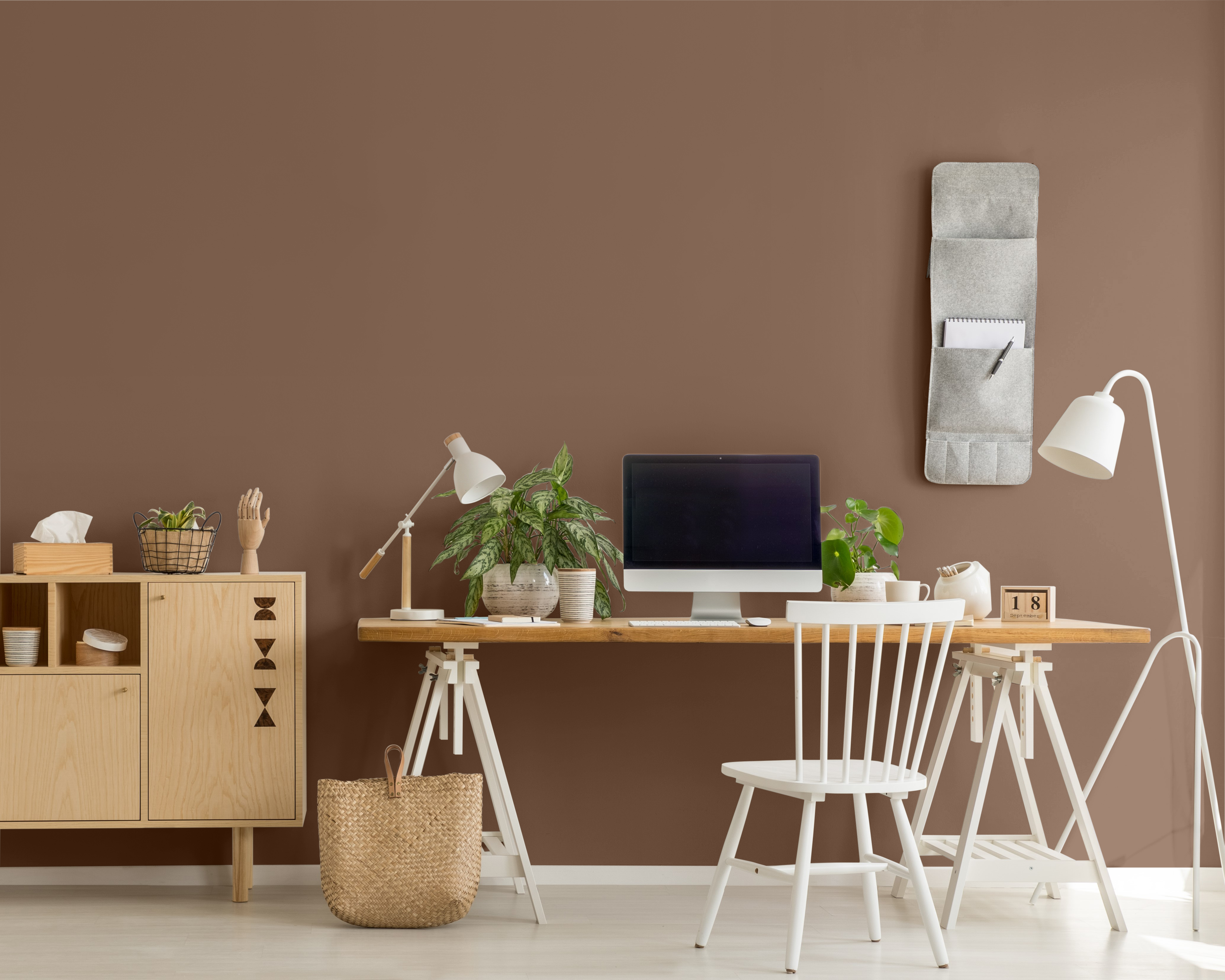 A photo of a wooden cupboard next to a desk with desktop computer and plants in home office interior. The wall color is painted in brown color called Wild Mustang. 