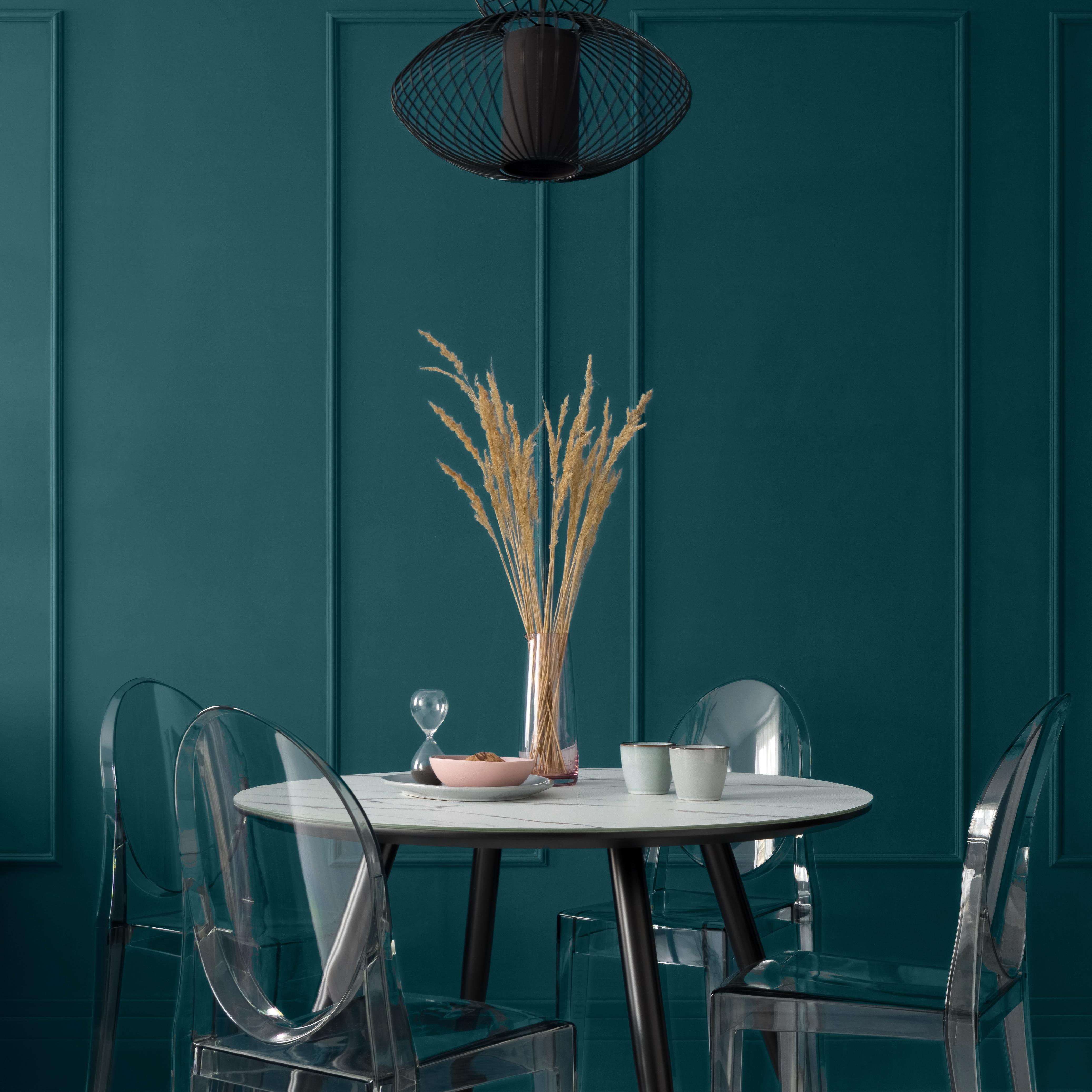 Fancy dining room with blue-green wall with molding and with modern table with black legs and marble style countertop, nice table decorations, four, new plastic chairs under black pendant lamp. The wall being featured is was painted in a deep teal color called Ocean Abyss. 