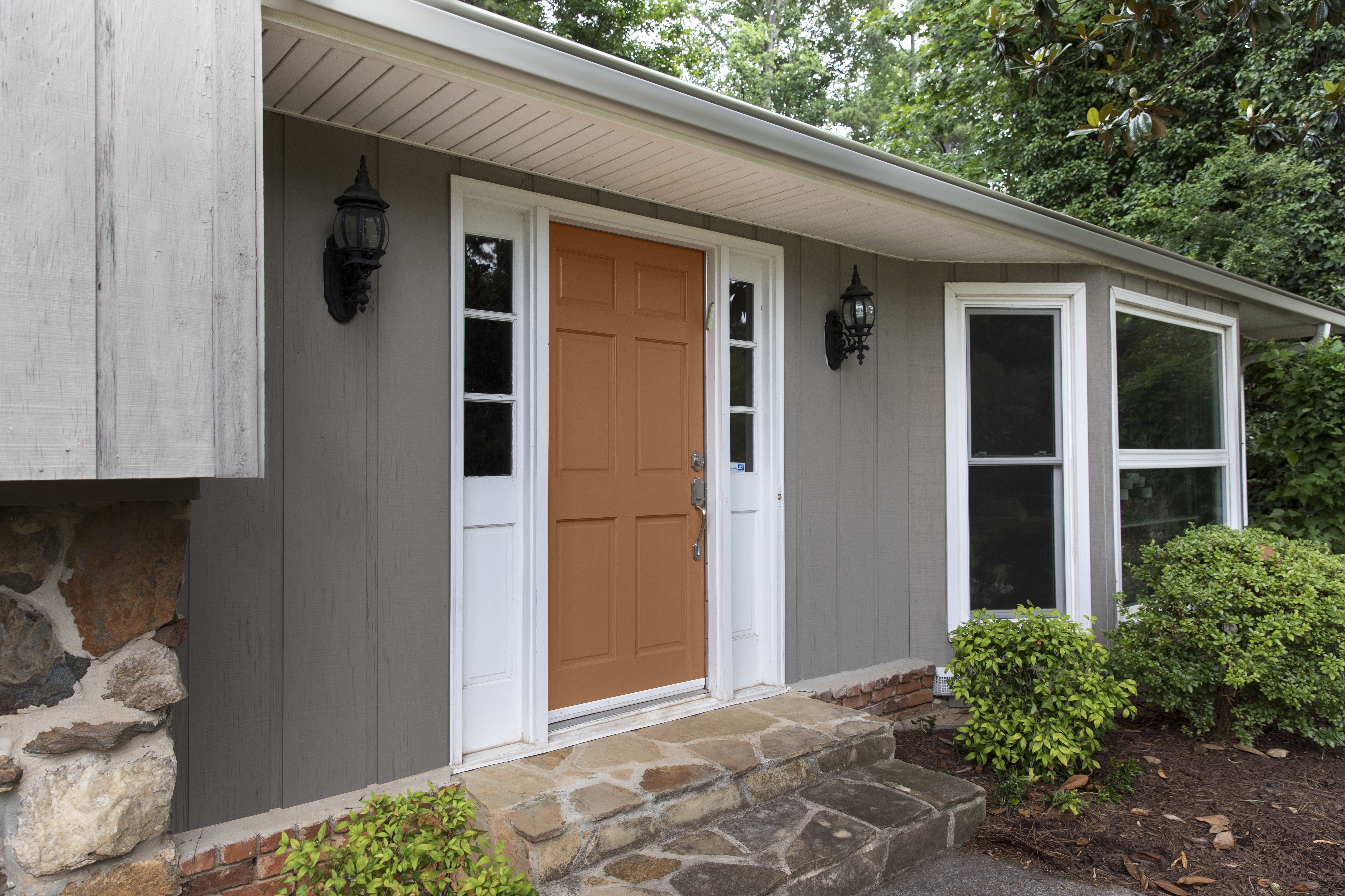 A mid range image of the front of a single family home.  The body of the house is painted in a mid-to-dark green with a green undertone called, Barnwood Gray.  The trim is featuring a super white color called ULTRA PURE WHITE.  The front main door features an inviting orange tone called Maple Glaze. 