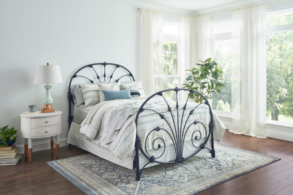 A bedroom with walls painted in a light blue color. Next to the bed is a white painted nightstand with gold accent legs and a gold lamp place on top.