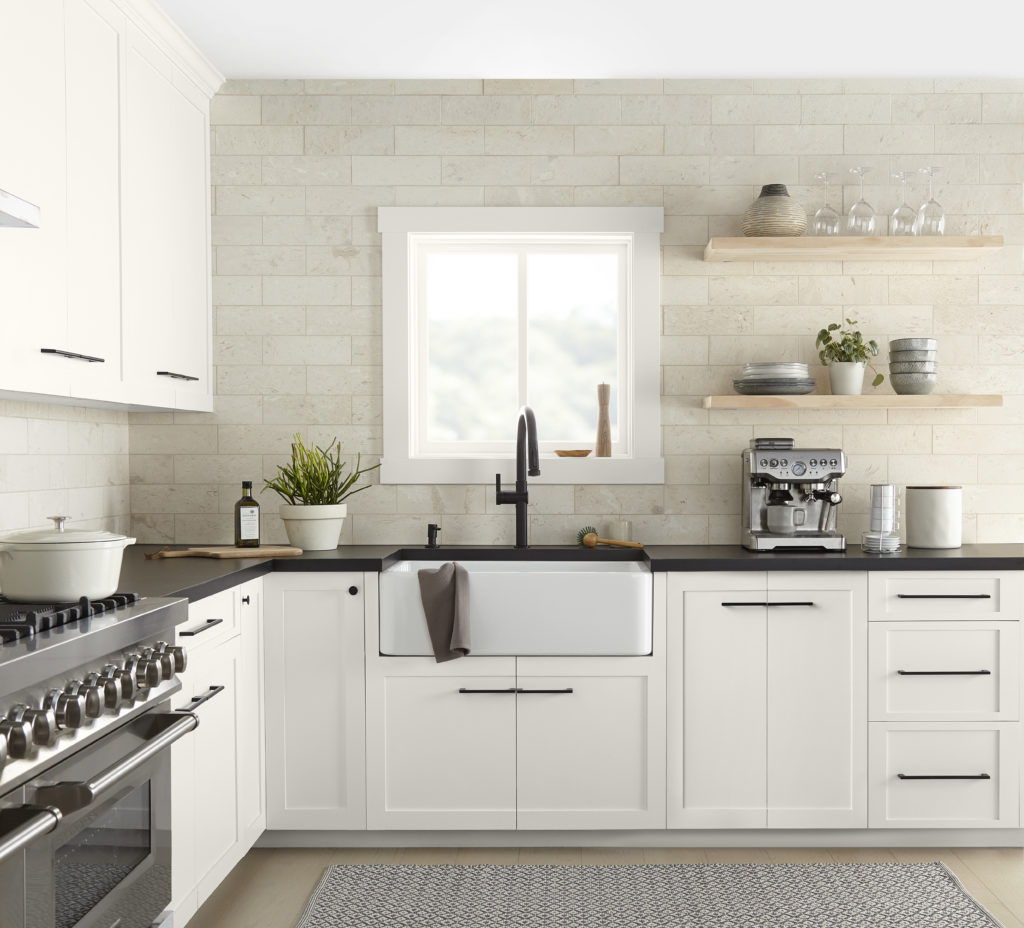 A modern farmhouse kitchen featuring white cabinetry painted with a warm white. The countertop is dark granite, and the backsplash is cream marble. The everyday essential serving ware can be on display on two natural wood free standing shelves.