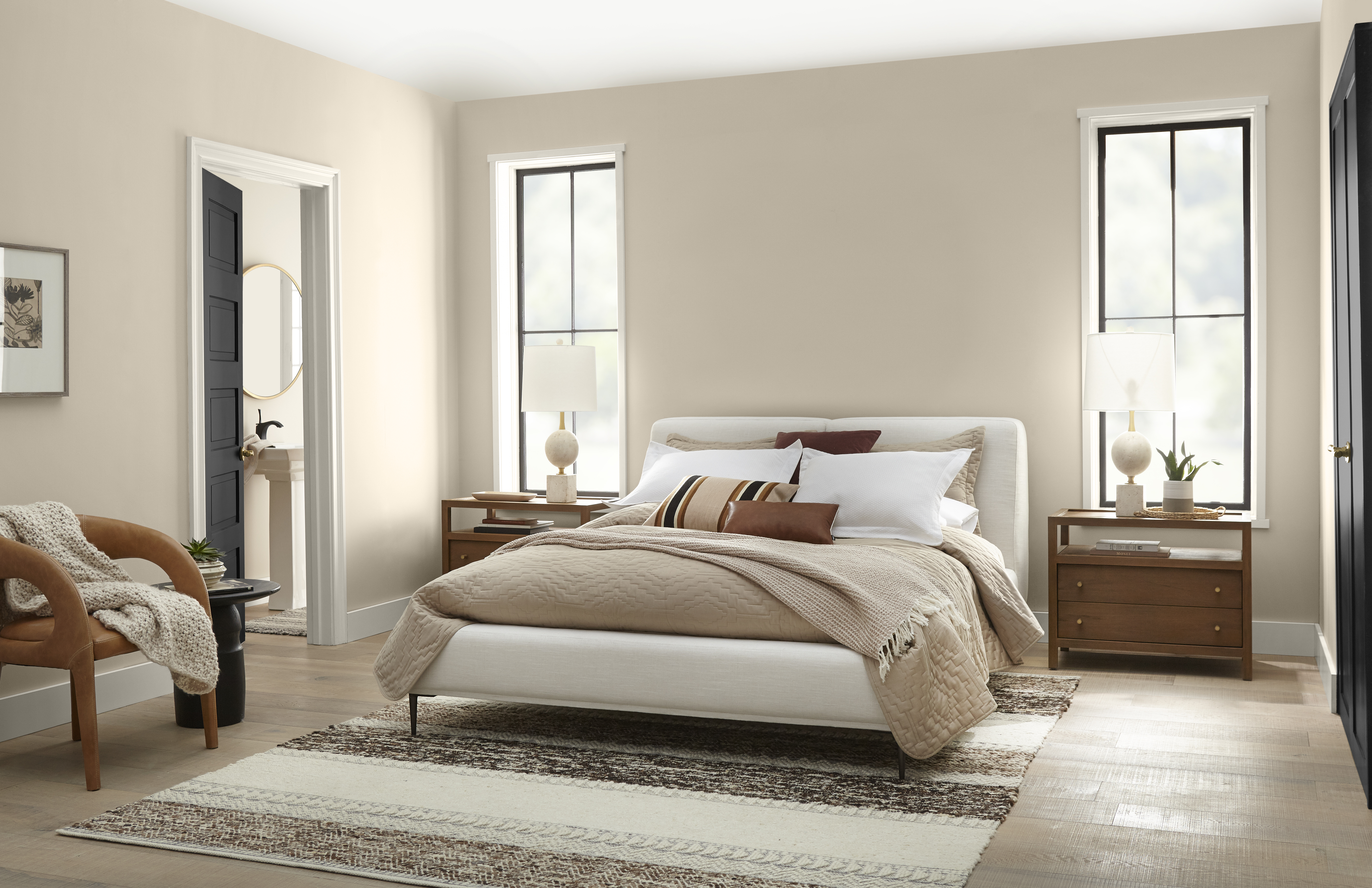 A bedroom painted in the colour Even Better Beige, with tall windows, modern neutral furniture, and black doors.