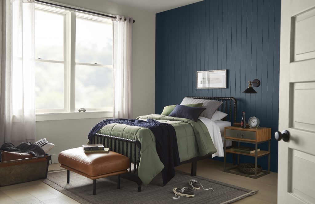 A charming bedroom with walls painted in Tranquil Gray and an accent wall painted in Midnight Blue, styled with deep greens, browns, and white in the décor and furniture