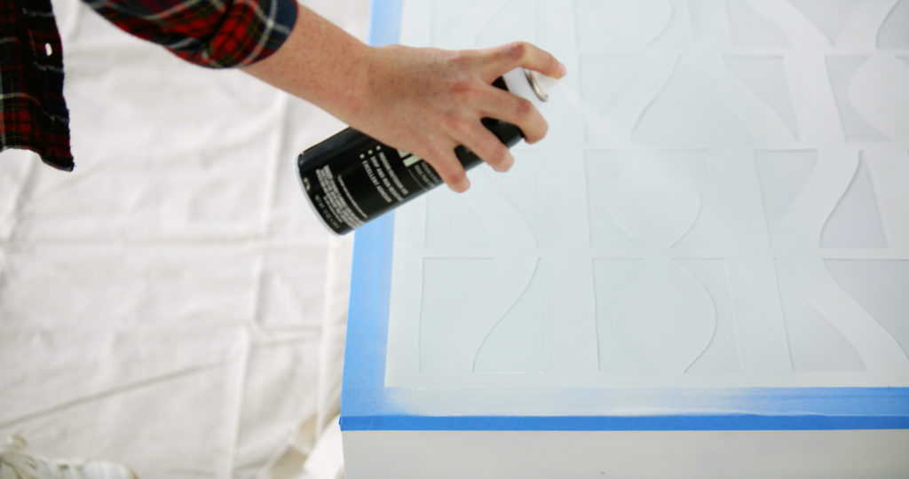 A person spray painting the top of a table that has a stencil taped to it