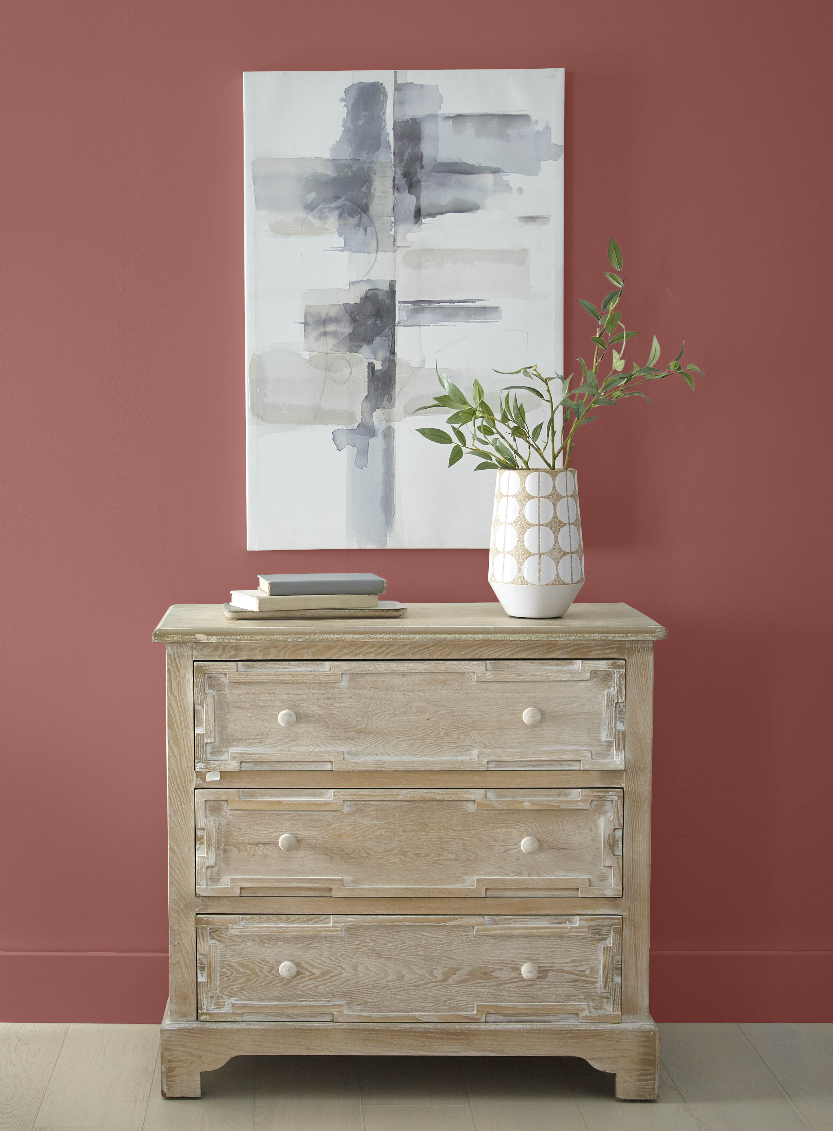A closeup of a wall in colour Vermilion, styled with a wooden chest of drawers and a neutral painting 