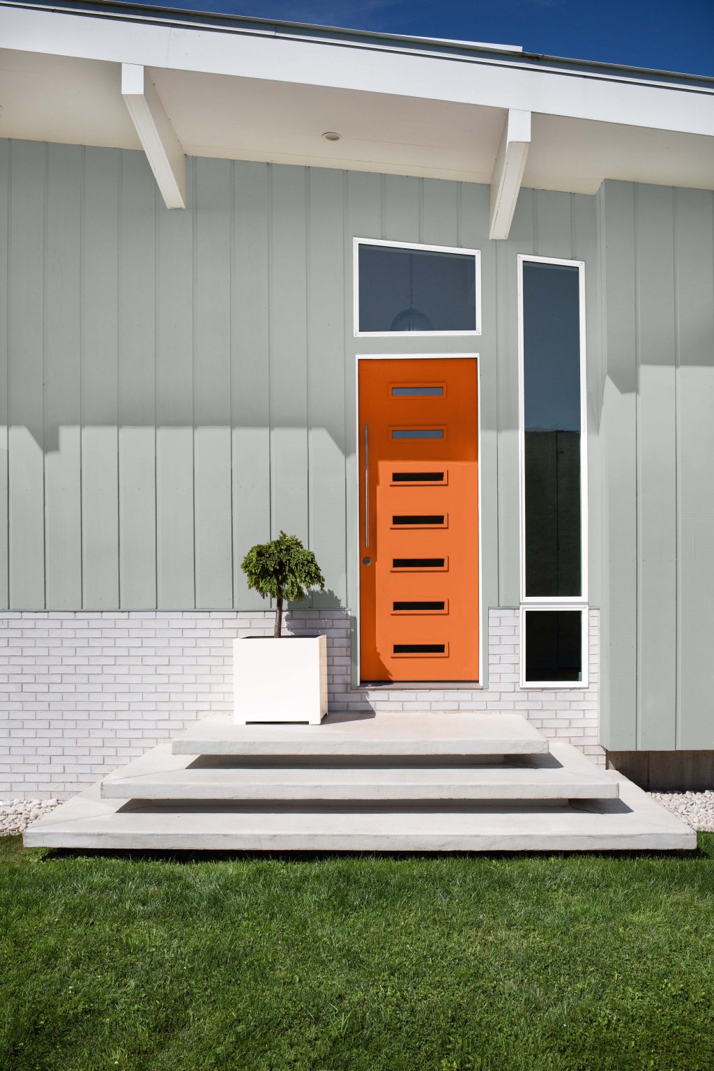 The exterior of a house with greyish blue siding, white trim, and the front door painted in the colour Tart Orange