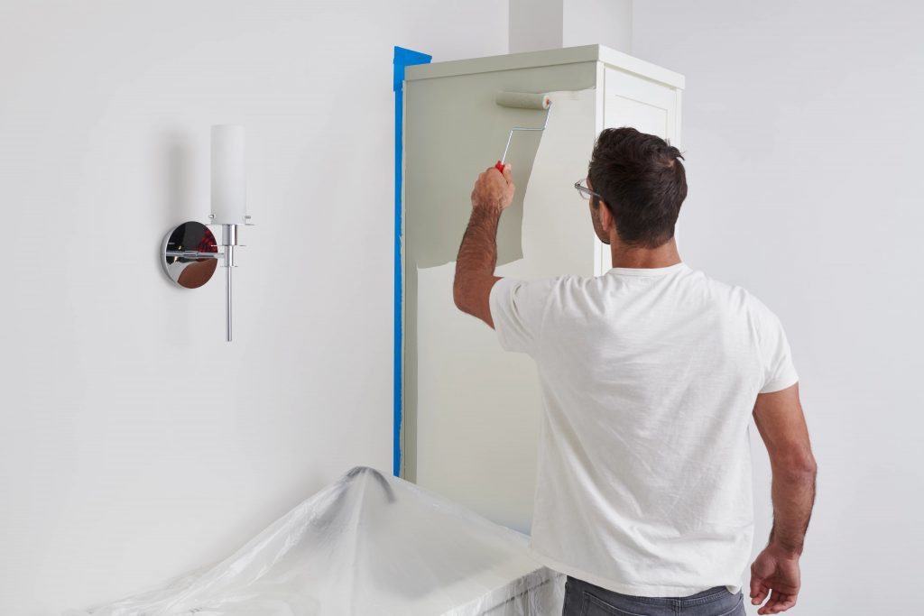 A man using a small roller to apply paint to the body of the bathroom cabinets 