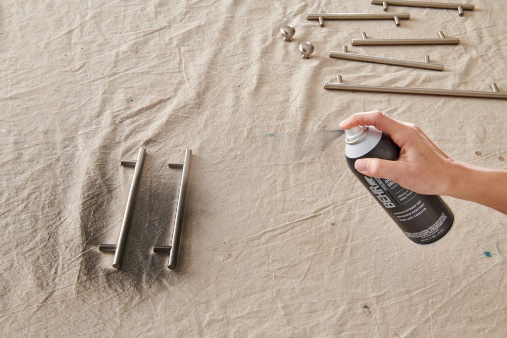 A closeup of a hand holding a spray paint can, applying spray paint to cabinet and drawer handles