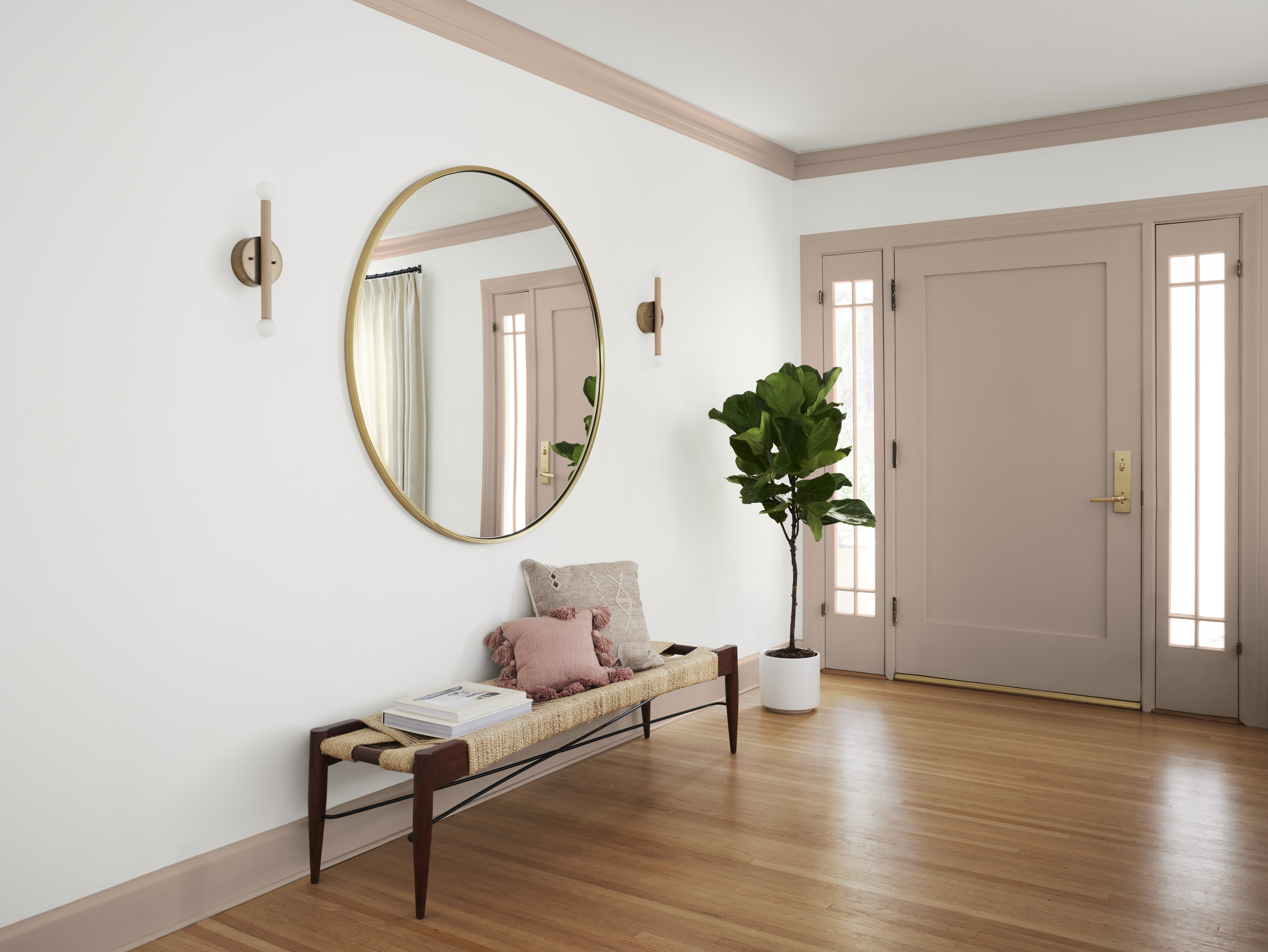A spacious entryway with white walls and the door/trim painted in a taupe colour, styled with a round mirror, a bench, and a plant