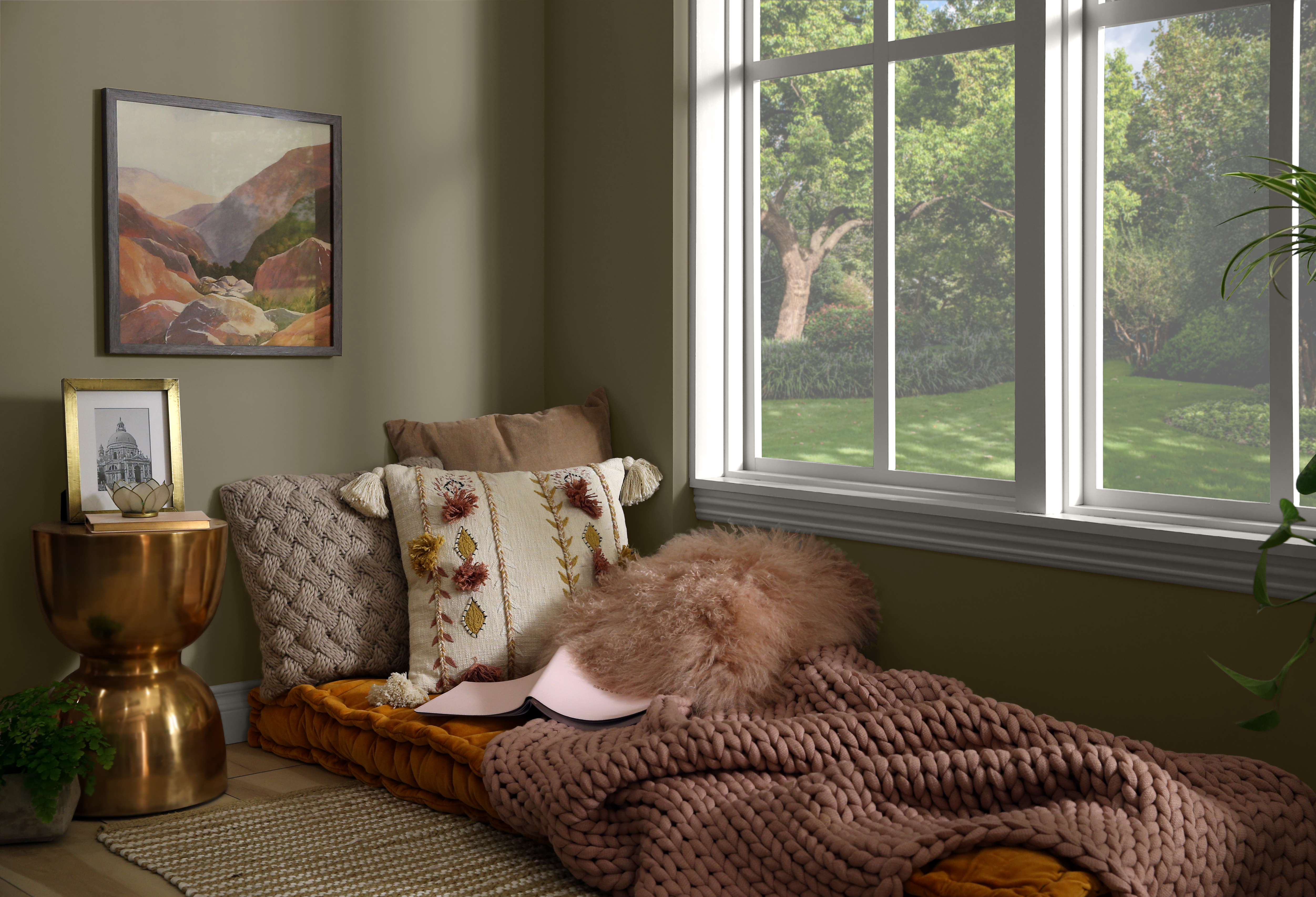 A cozy reading and lounging corner next to a large window and styled with pillows and blankets, with the walls painted in a dark olive-brown colour