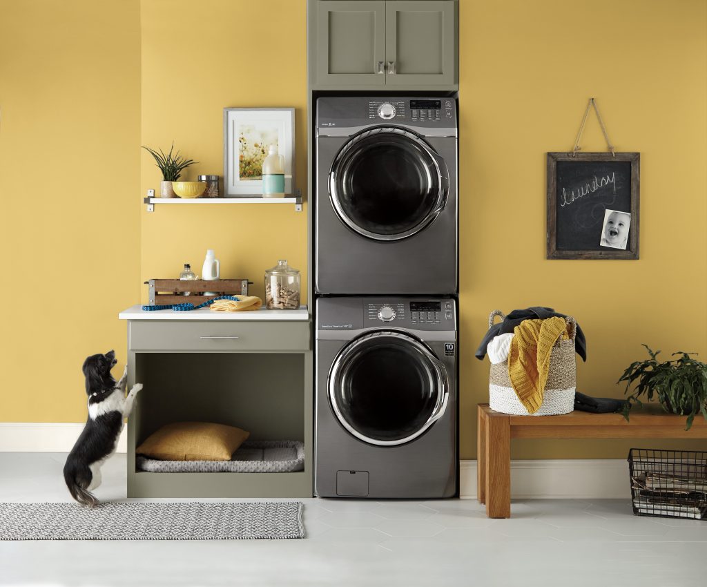 A spacious laundry room with walls painted in a bright yellow colour, with a stacked washer and dryer and wood bench
