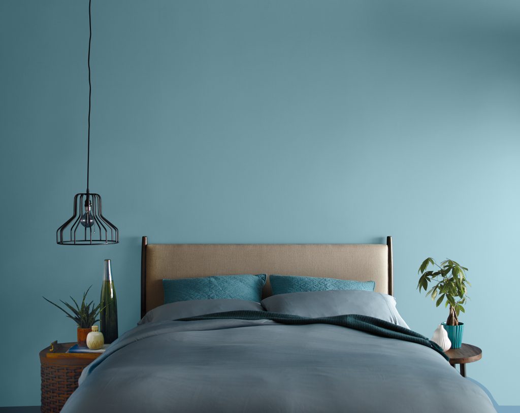 A bedroom with the headboard wall painted in a muted blue colour, styled with a neutral bedframe, blue linens and pillows, and side tables