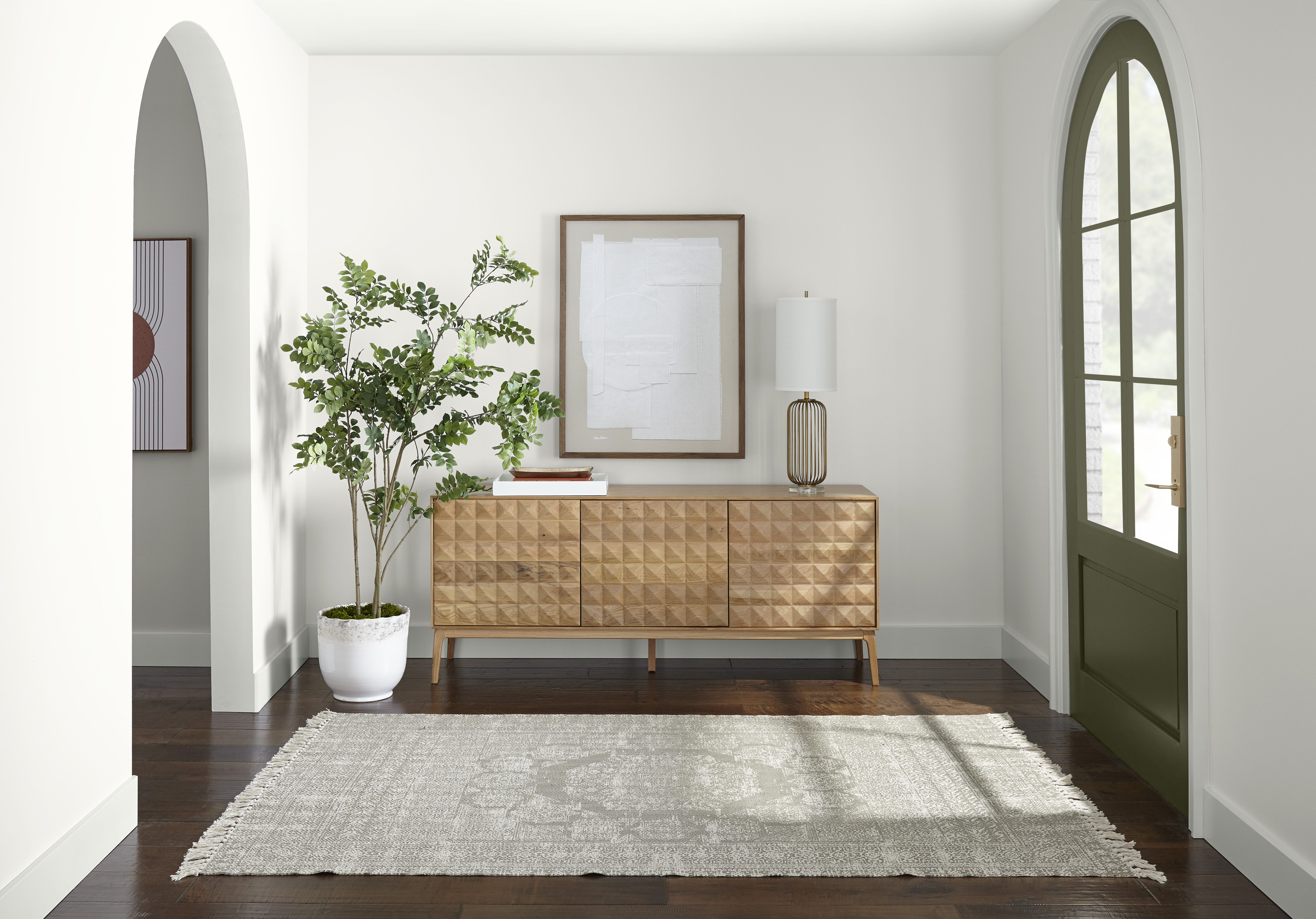 A spacious entryway with walls and trim painted in white, styled with a wood sideboard, large potted plant, a framed piece of artwork, and large grey rug 