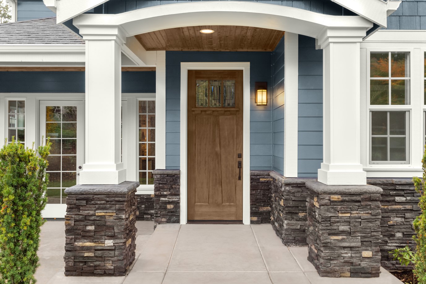 A closeup of the front of a house with blue siding, white trim, and a brown semi-transparent wood door and port ceiling