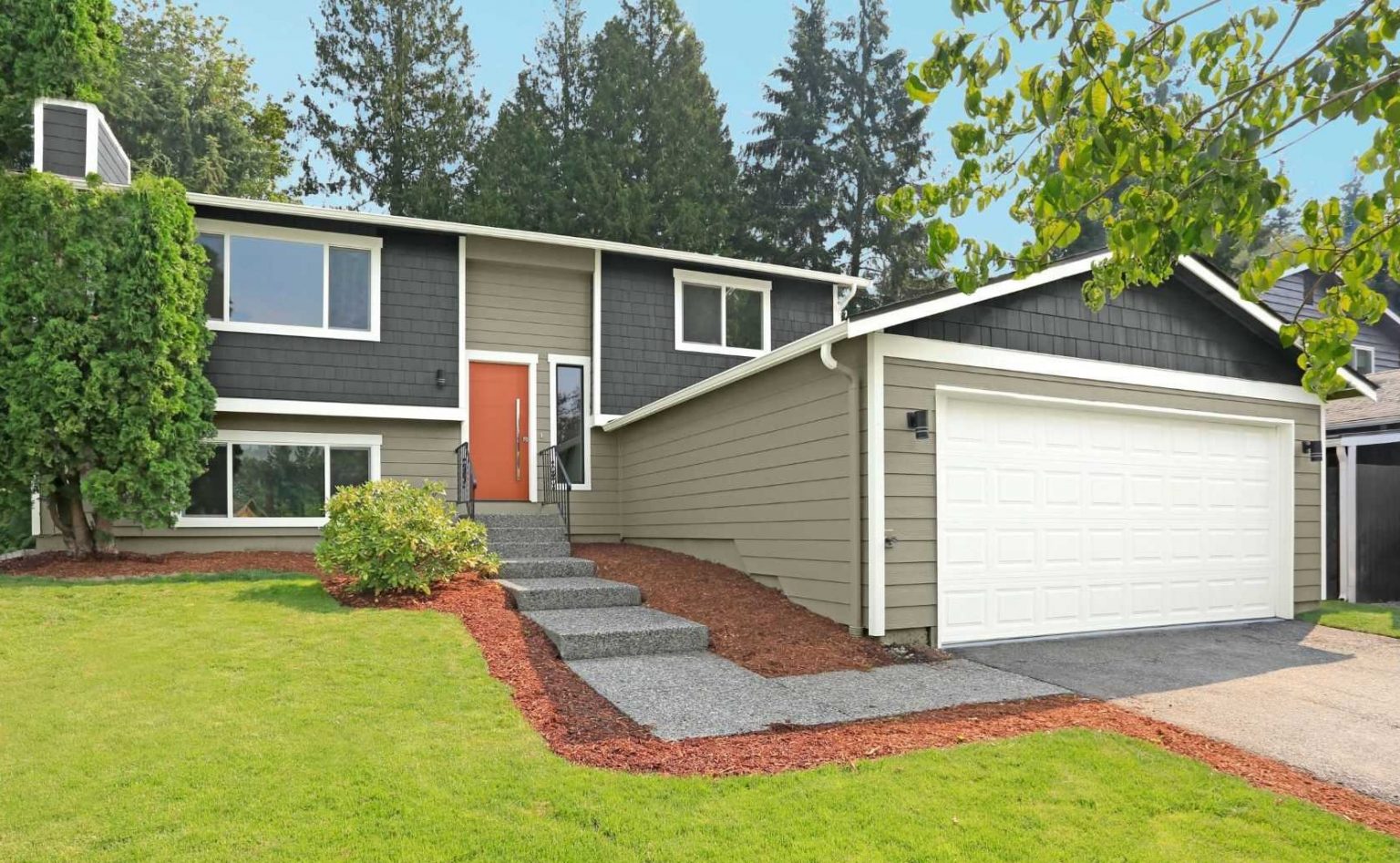 The exterior of a split-level Ranch style house with dark grey shingles and earthy green siding and a bright orange door