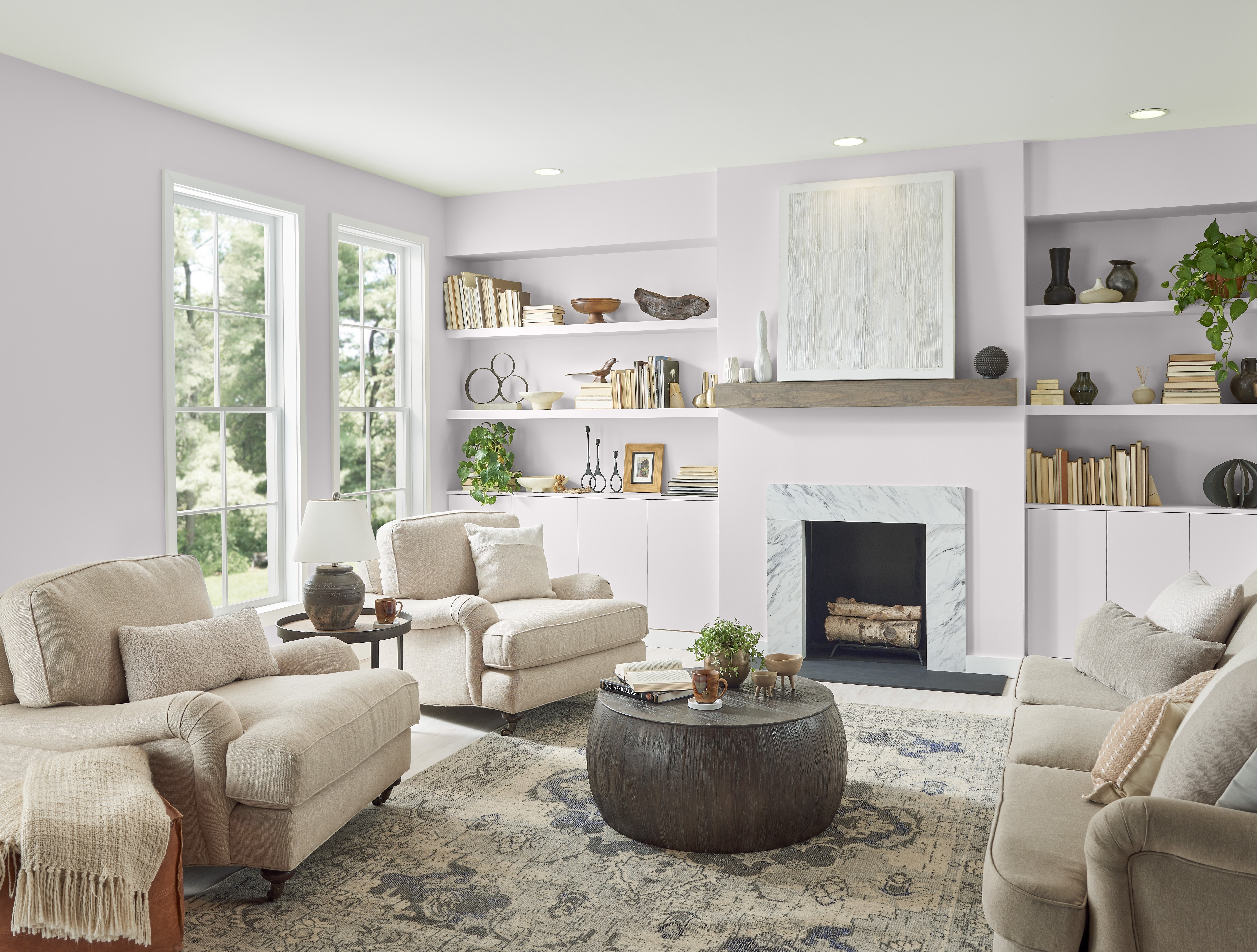 A bright and welcoming living room with an almost neutral colour palette. The room has large windows, allowing natural light to fill the space. A fireplace is present on the right side of the image, above which hangs an abstract piece of art.