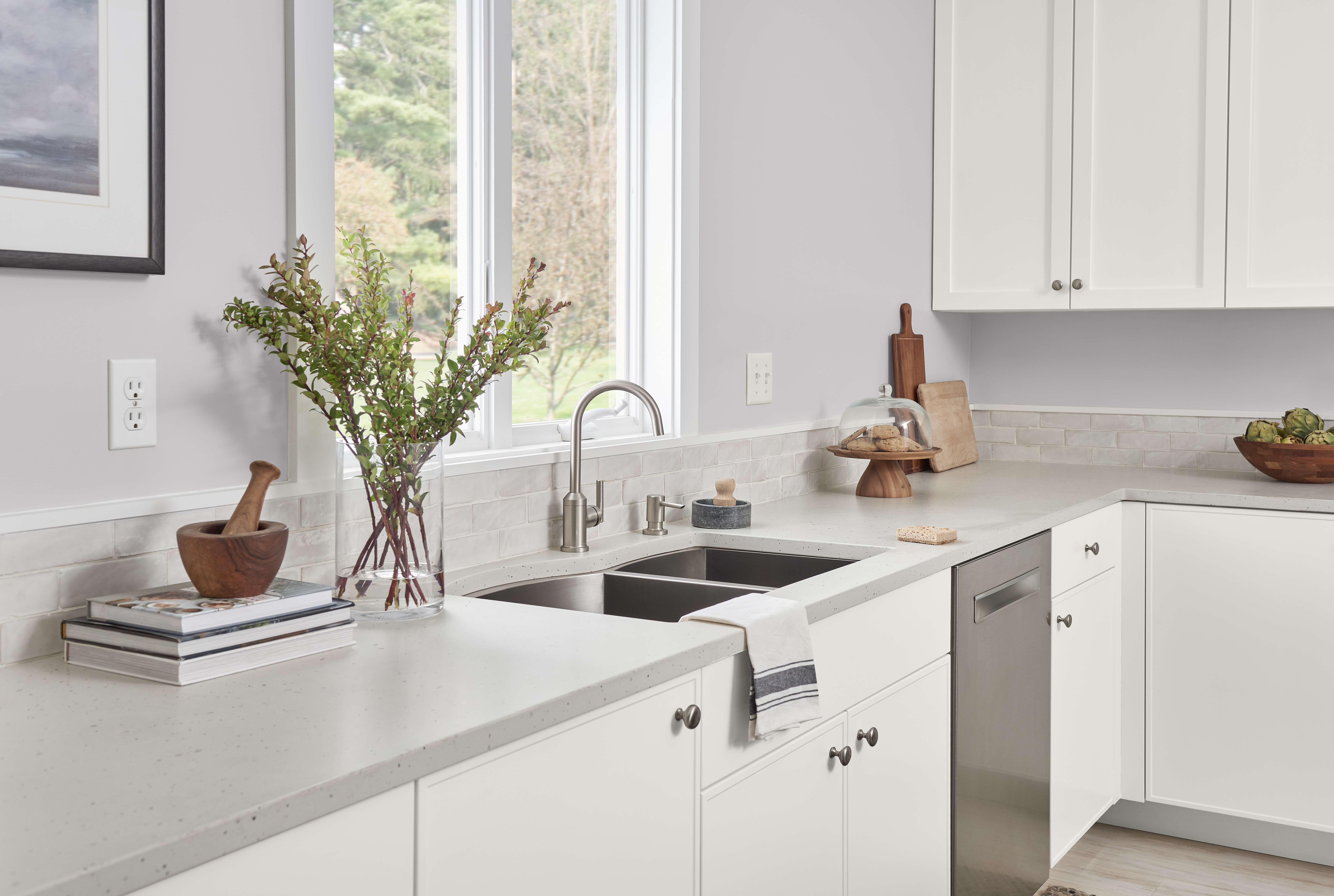 A kitchen with light grey-violet colour walls and white cabinetry features a white countertop with a built-in stainless-steel sink. The counter includes a wooden mortar and pestle, a stack of books, and a vase with green foliage. To the right of the sink, there are cutting boards, a cake stand with pastries, and a bowl of fruit. Large windows allow natural light to fill the space.