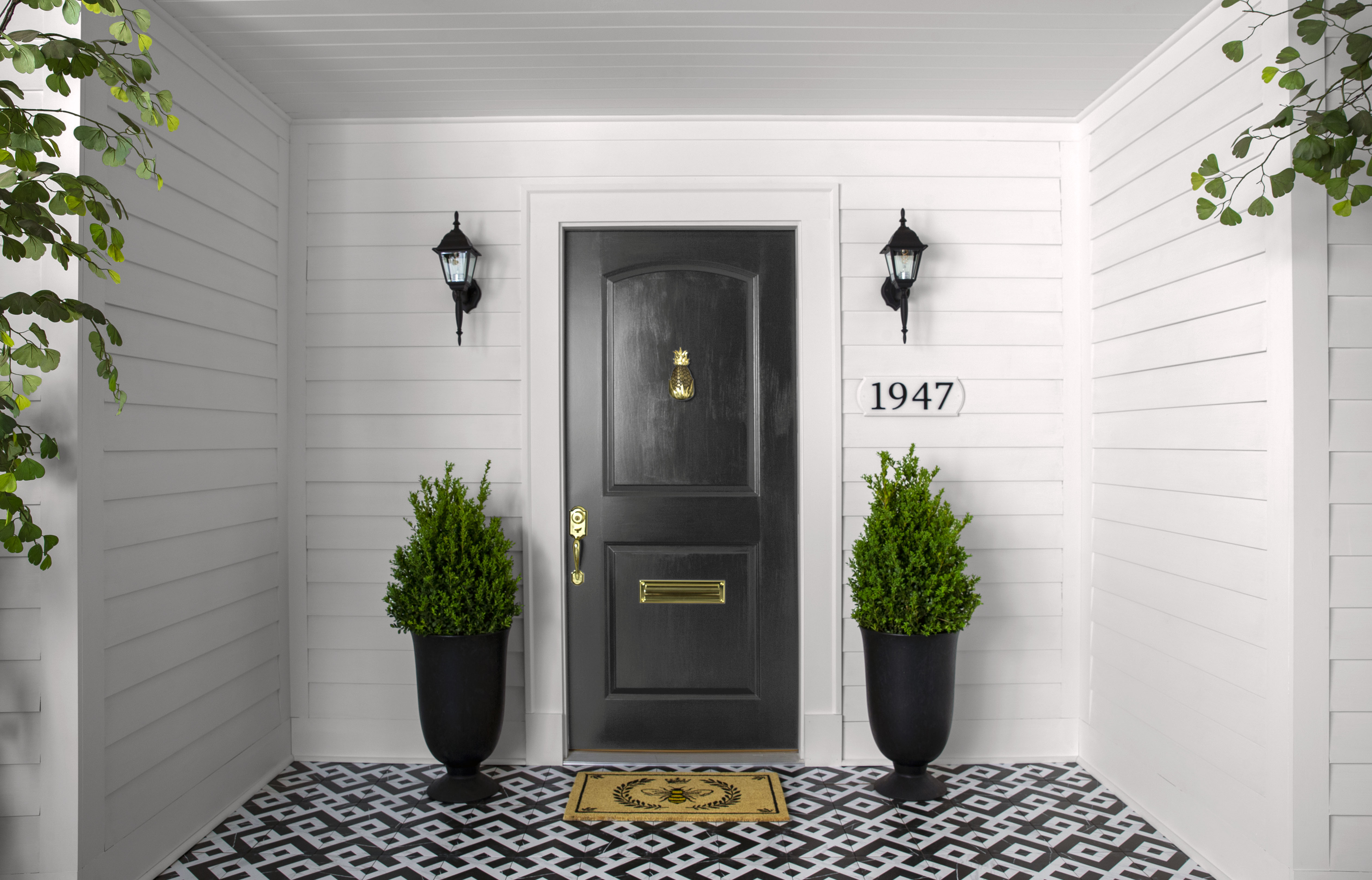 A house facade featuring a light purple, grey exterior with a black front door centered between two tall planters holding lush greenery.