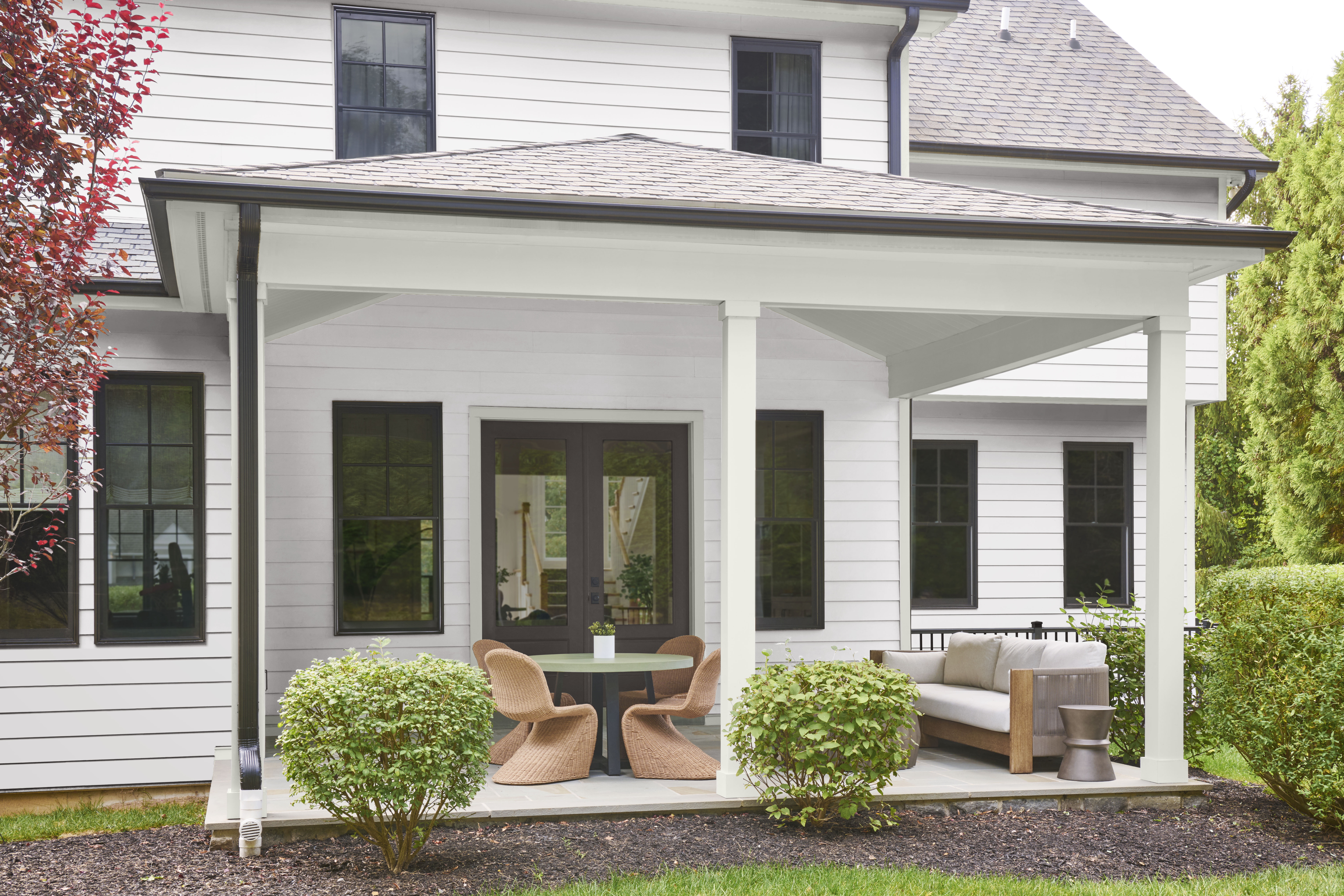 A two-story stucco house painted light grey (Rock Crystal) with white inset trim and patio ceiling, and soft black accent trim. Patio features outdoor furniture including a sofa, round table and sculptural chairs made with natural material and colour.