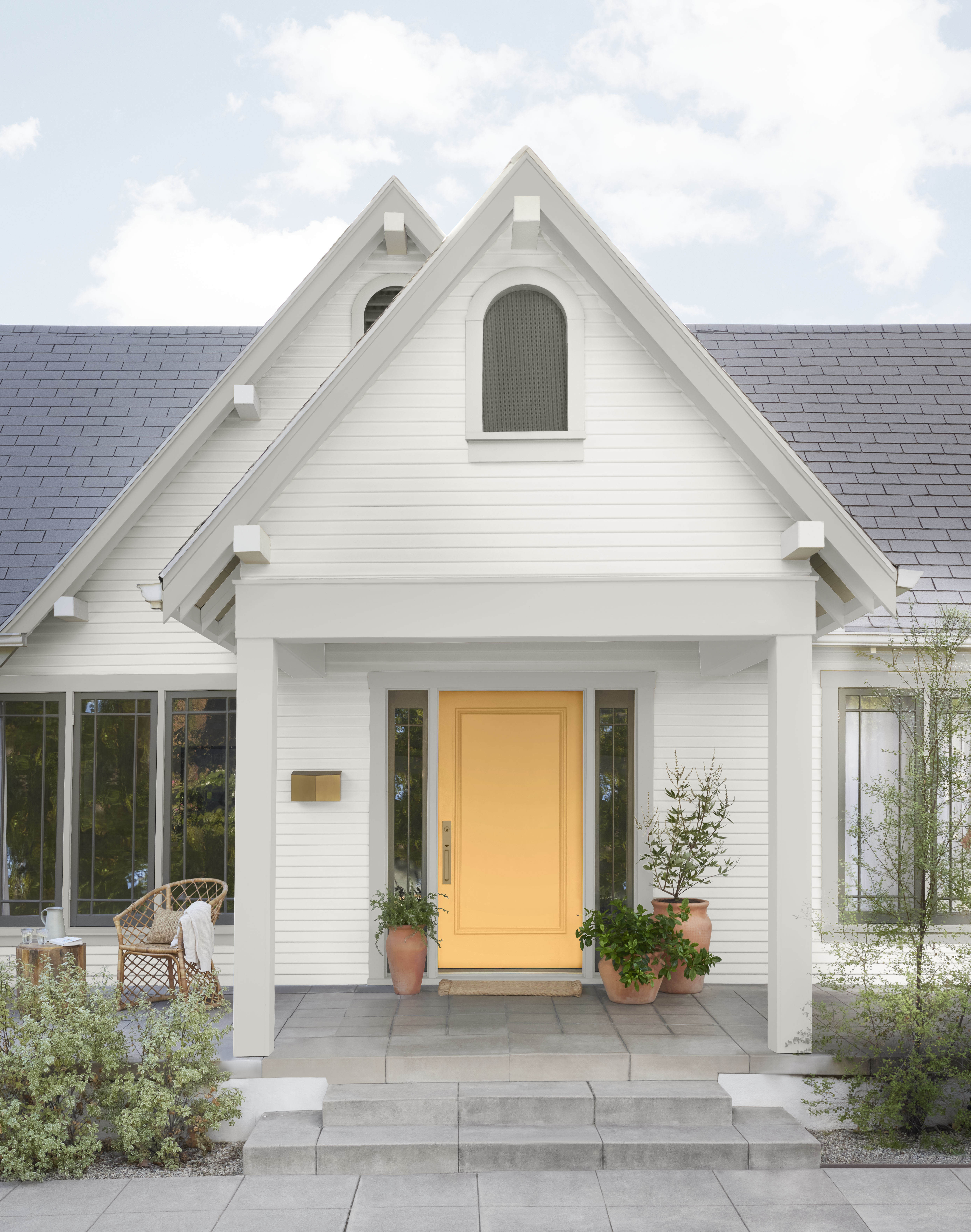 A Tudor-style influenced home exterior with a double-pitched roof, painted off-white with trim in a muted green-gray shade called Historical Gray. A bold yellow front door adds a contemporary pop of brightness, blending classic architecture with fresh, modern color accents.