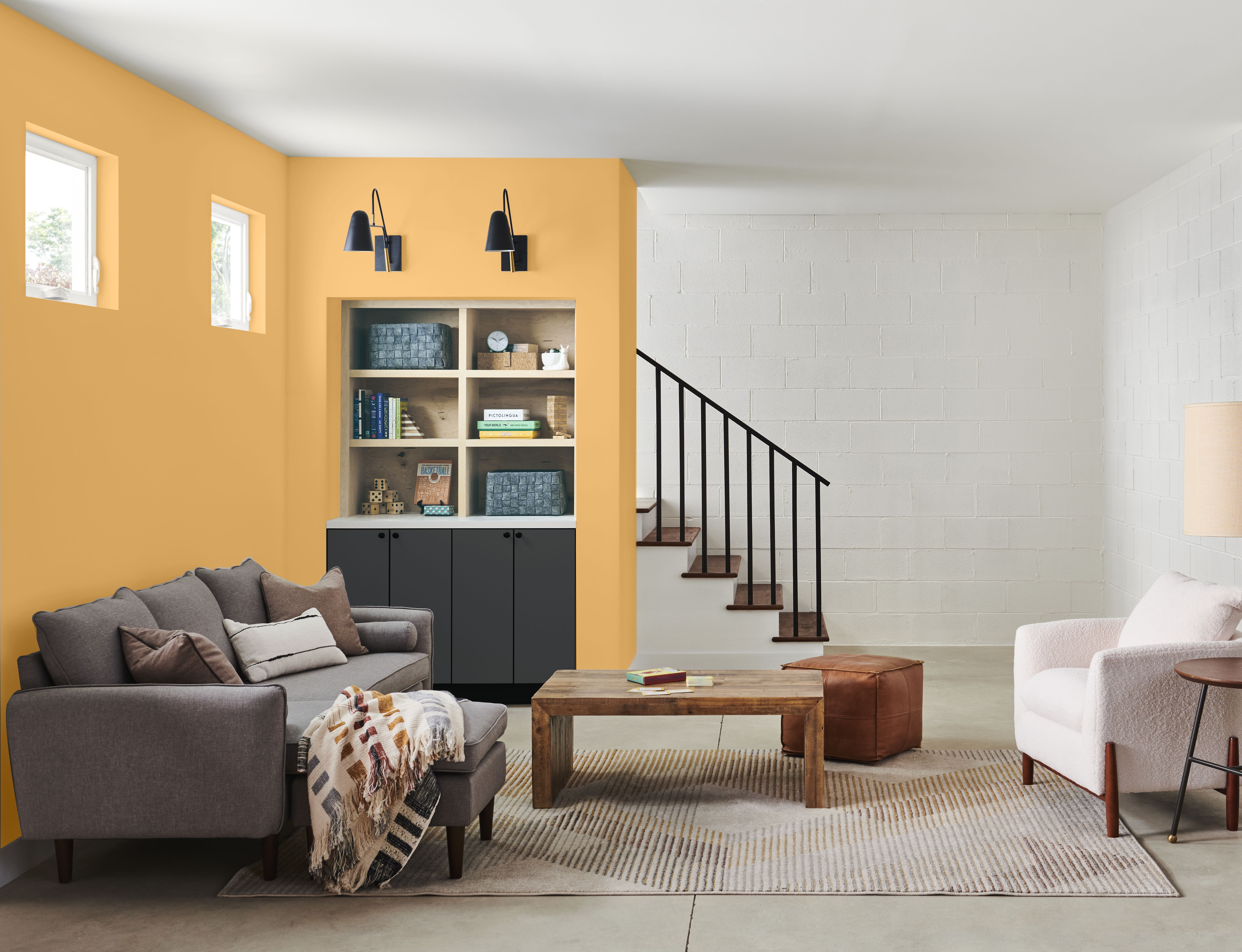 A basement living space featuring a comfortable sofa, an L-shaped accent wall painted in a vibrant yellow, and built-in shelving with lower cabinets painted in a soft black, creating a cozy yet modern atmosphere with bold colour contrast.