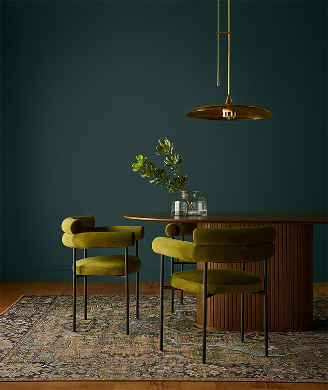 A Modern luxe dining room featuring a rich wooden oval table and two retro-modern velvet chairs. The wall is painted in Hidden Gem, a smoky jade green that whispers elegance and speaks volumes in sophistication.
