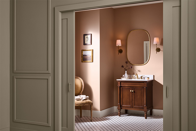 An elegant bathroom with warm light warm pink-coloured walls, a wooden vanity with white countertop, and a gold-framed oval mirror. Two wall sconces with peach shades flank the mirror, and a small chair with folded towels sits against the side wall beneath framed artwork.