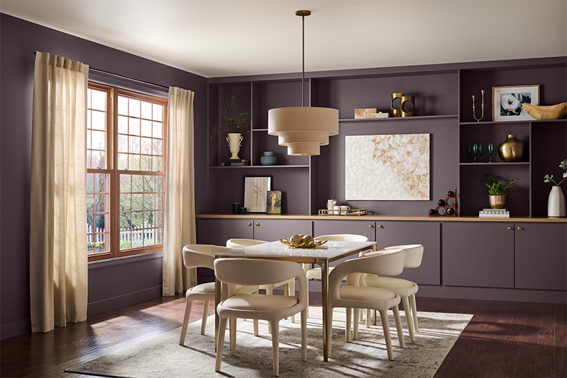 An eclectic dining room with dark purple walls and a large window with beige curtains. The room features a white dining table surrounded by off-white chairs on a light-coloured rug. Above the table hangs a beige, tiered pendant light. The back wall has built-in shelves and cabinets, decorated with various items including vases, books, plants, and artwork.