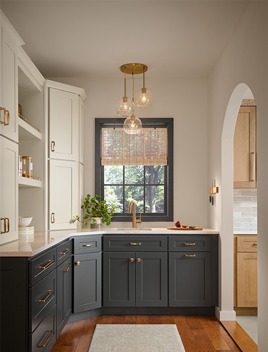 A beautiful kitchen pantry with two-tone cabinetry featuring dark grey lower cabinets and light beige upper cabinets, accented with brass hardware. A black-framed window above the sink has woven blinds, with a brass faucet below.