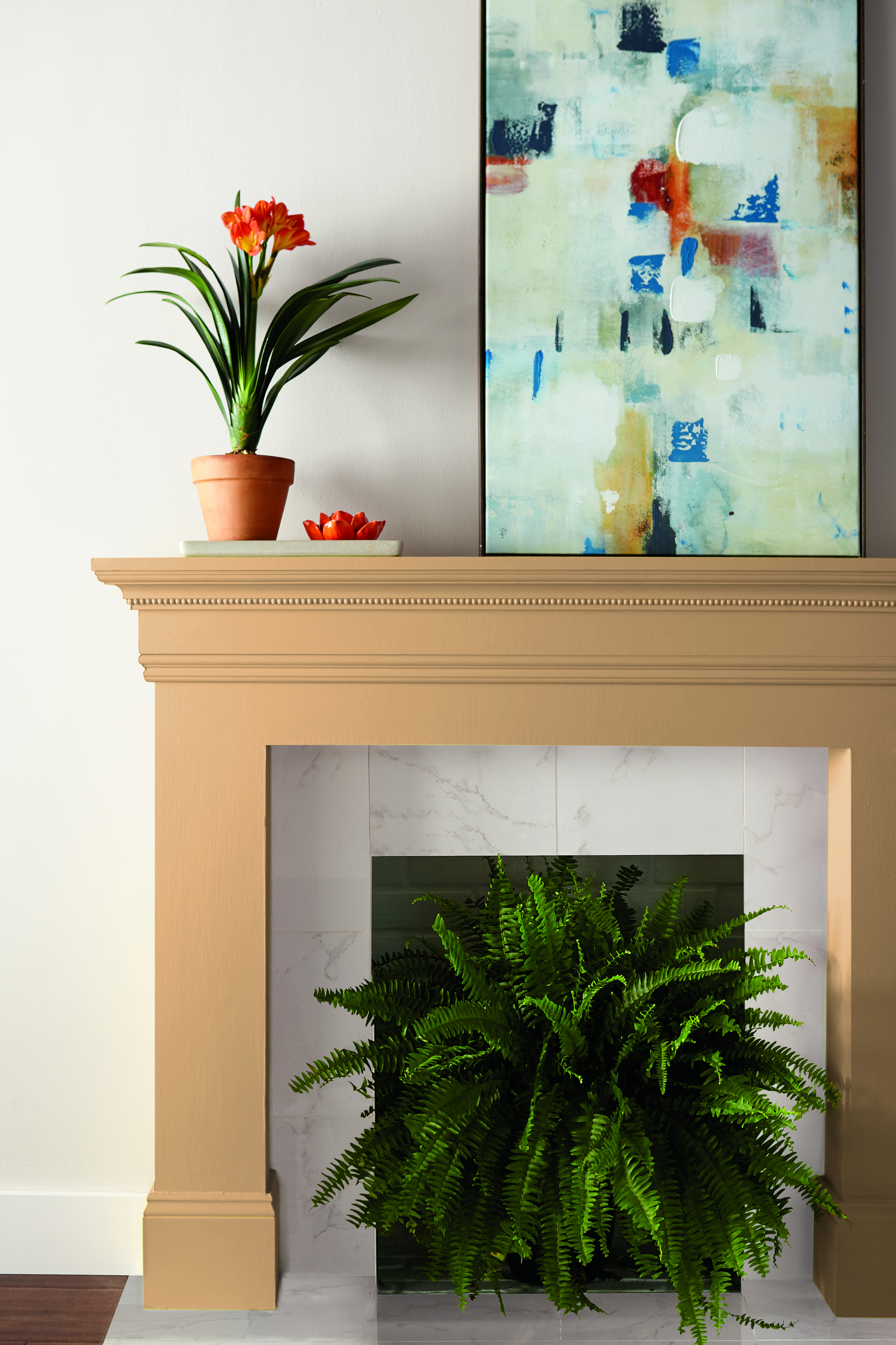 Golden, yellow-painted fireplace mantel with orange flowers, abstract art, and a green fern inside; white wall background.