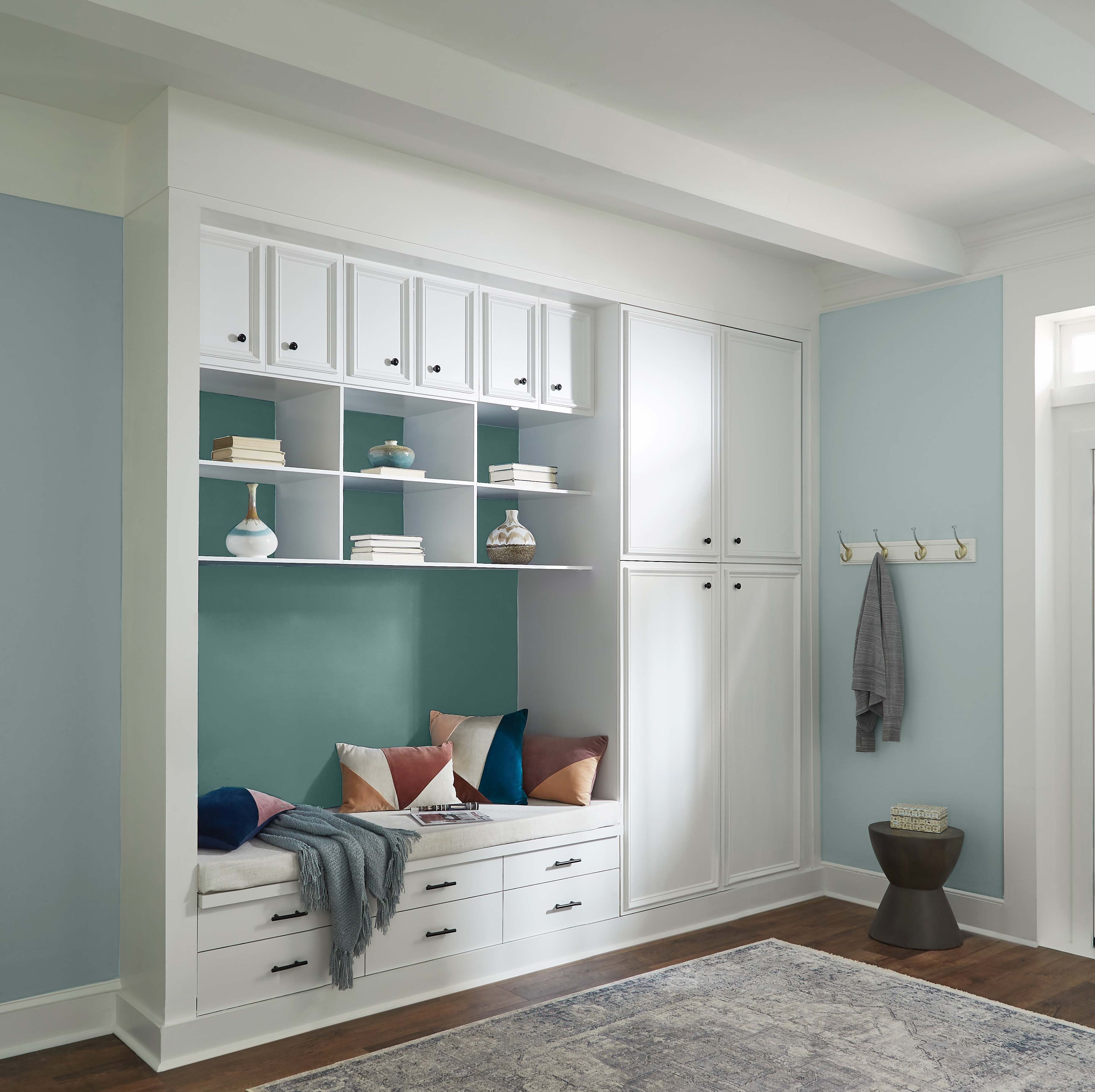 A hallway-entry area with built-in storage nook surrounded by white cabinetry and shelving. The nook features a dark blue accent wall behind the shelves and bench. The surrounding walls are painted in Watery. The bench is styled with colourful throw pillows and a blanket.