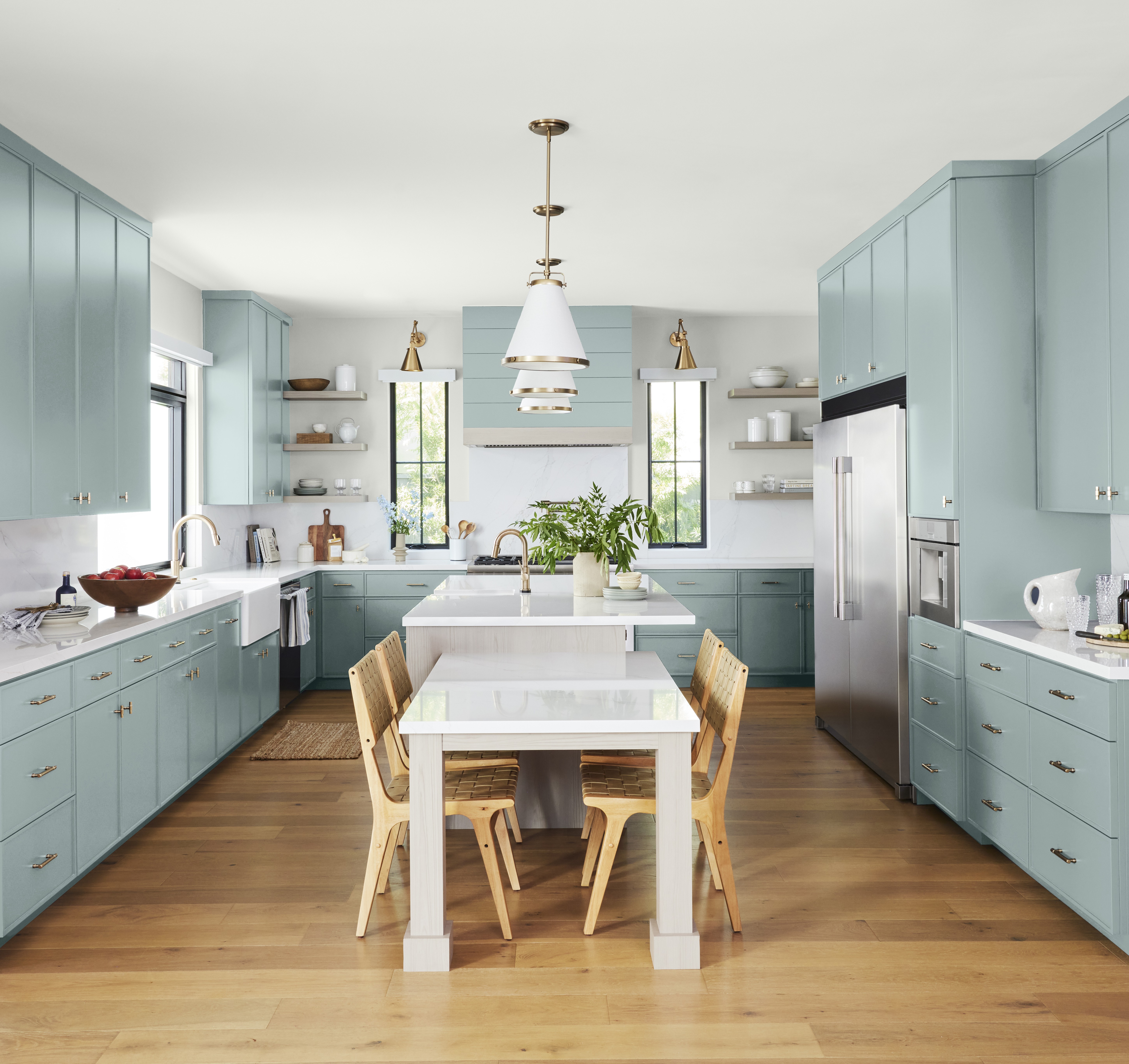 A spacious kitchen with cabinets painted in a soft blue-green tone called Watery. The room features a white island, a white eating area and light wood dining chairs. White coloured backsplash and countertops and warm wood flooring contrasts beautifully with the cool-toned cabinetry.