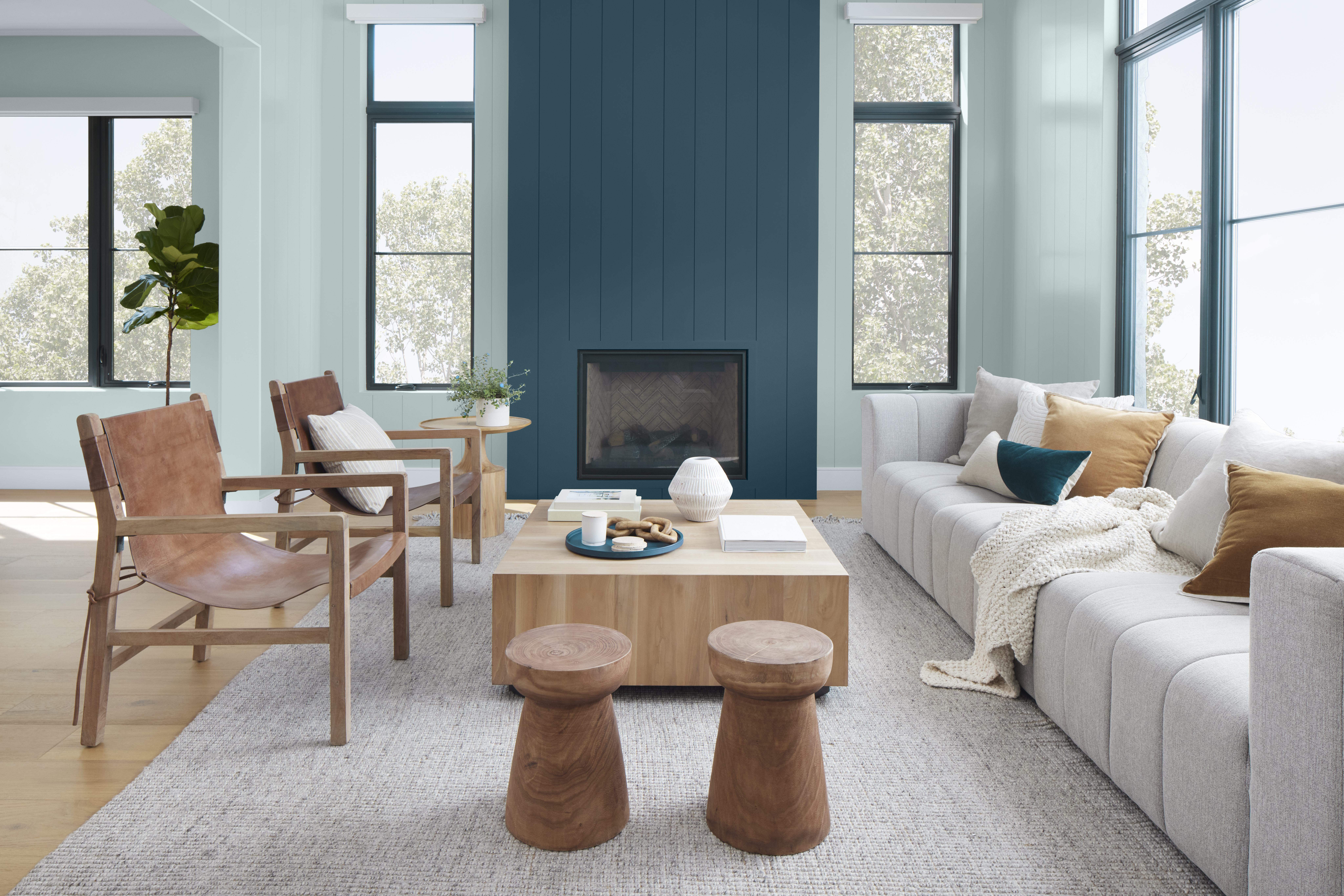A modern living room with a lot of natural light and painted with Watery. It features a tall shiplap fireplace wall painted in a deep blue shade called Nocturne Blue. The space features a light grey sofa, a wooden armchair with a tan cushion, and a square wooden coffee table with decorative objects.
