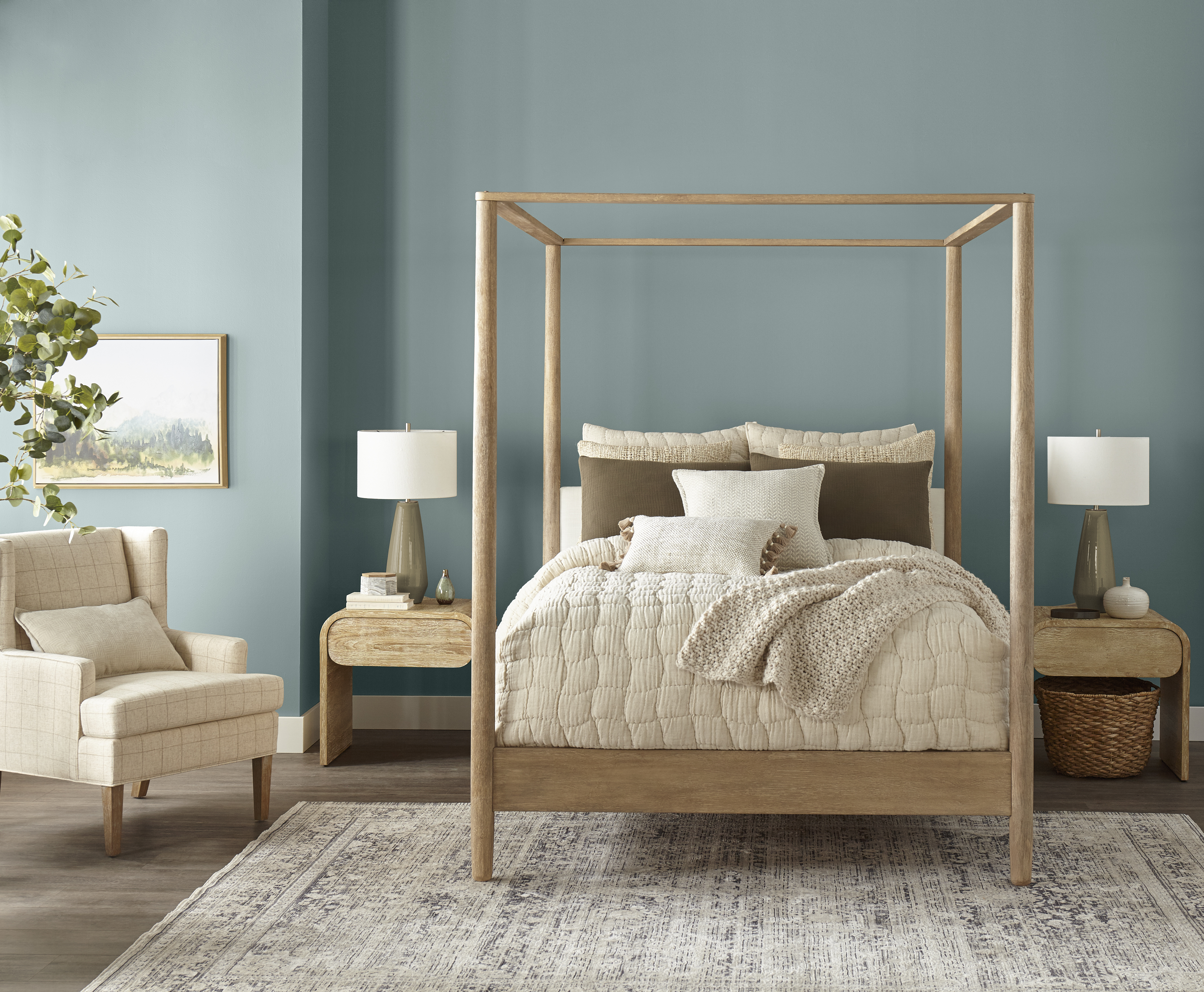 Cozy bedroom featuring a four-poster wooden bed with layered neutral bedding, including a quilted comforter and textured throw blanket. The walls are painted in a soft light blue, creating a calm and balanced atmosphere. Two wooden nightstands with lamps flank the bed, complemented by a light armchair, a patterned area rug and a potted plant for greenery.