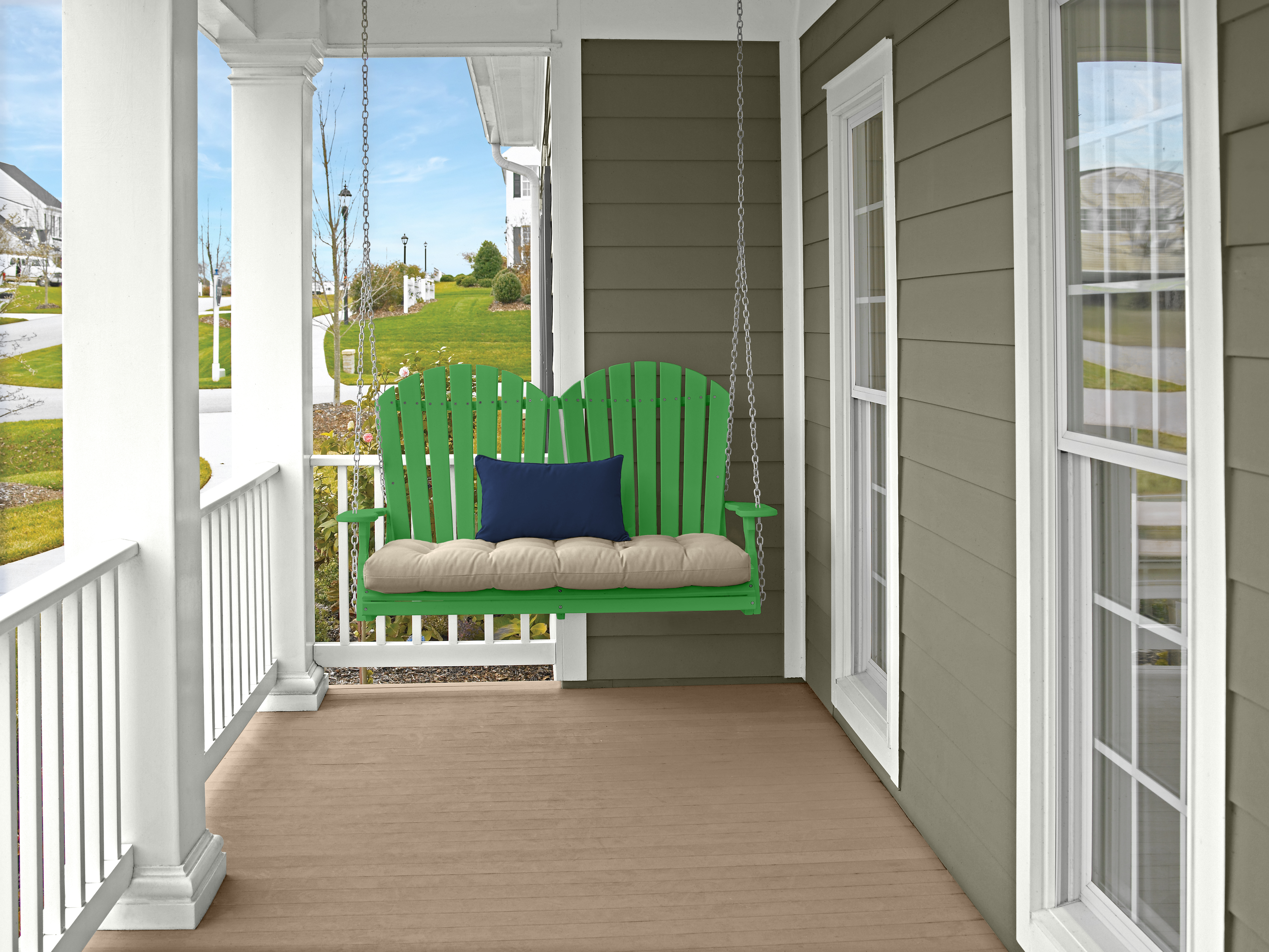 A front porch with white columns and railing, green porch swing with beige cushion and navy blue pillow, light wood flooring, and muted green colour house siding overlooking a grassy neighbourhood. The porch wood stain colour is Taupe and the finish a semi-trasparecent opacity. 