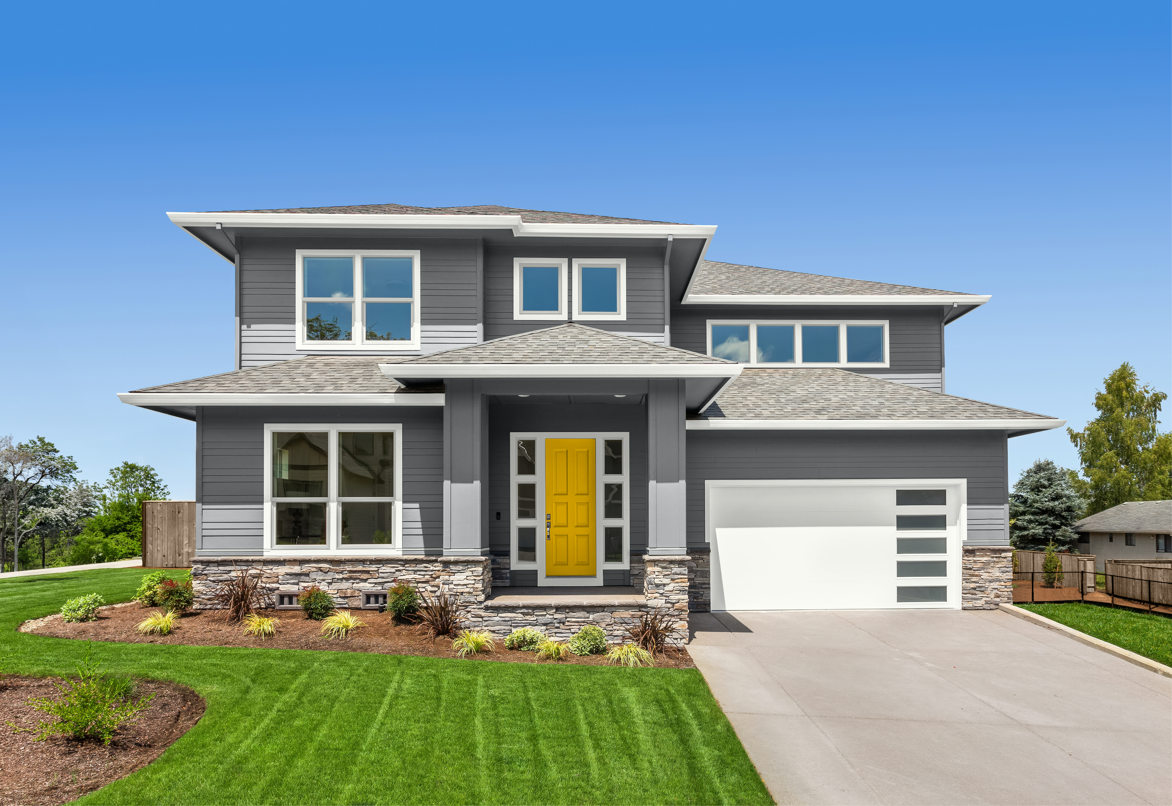 A new construction two-story grey house with a bright yellow front door, white trim, and a covered entry supported by square columns. The home features large windows, a stone-accent base, and an attached white garage under a clear blue sky. The wood fence has been finished with the 2026 BEHR wood stain colour of the year, Taupe, in a semi-transparent finish.  