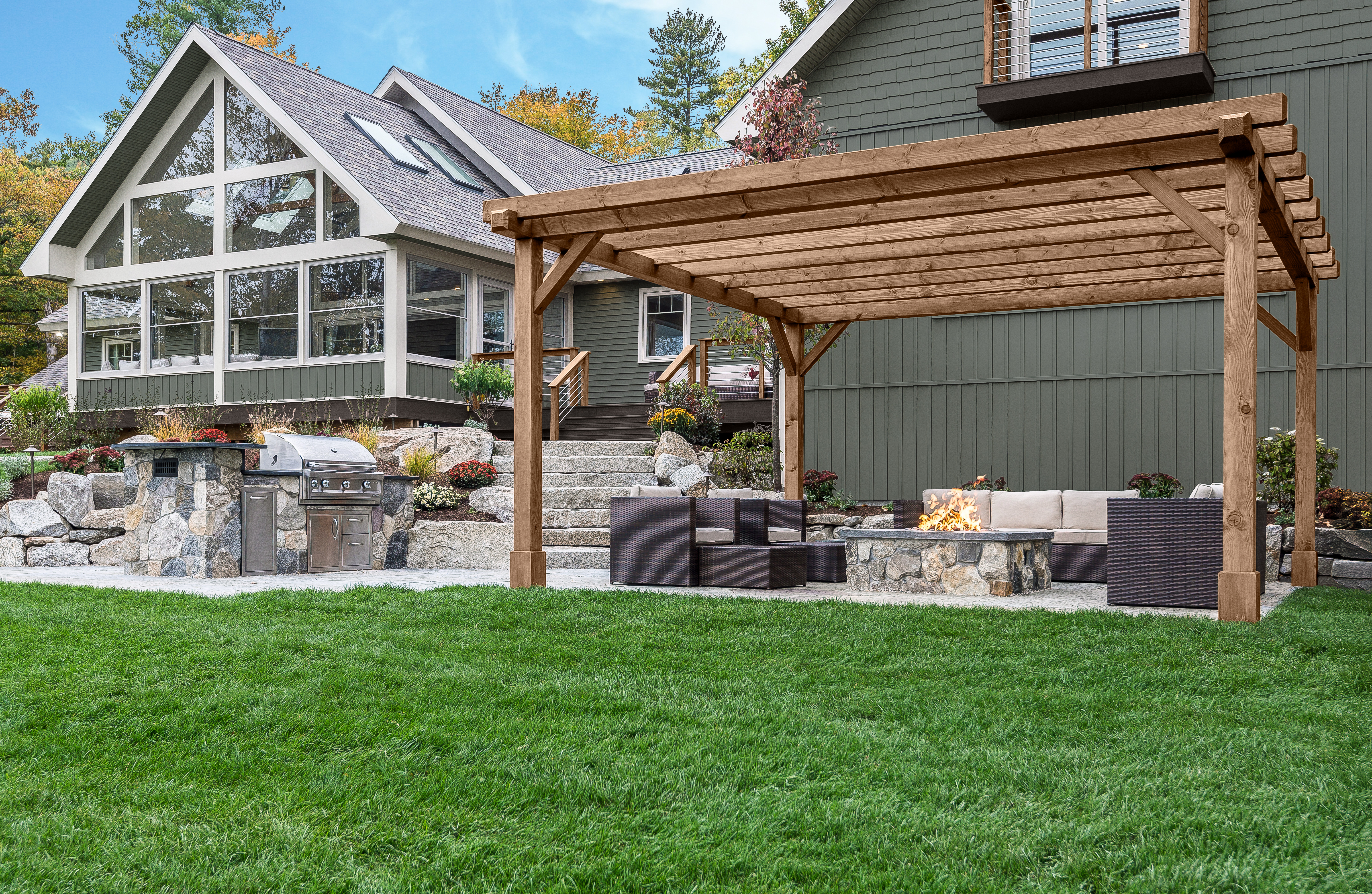 A backyard patio with a wood pergola stained with Taupe, outdoor seating around a fire pit, and a grill area beside a modern home with large windows and landscaped yard. 