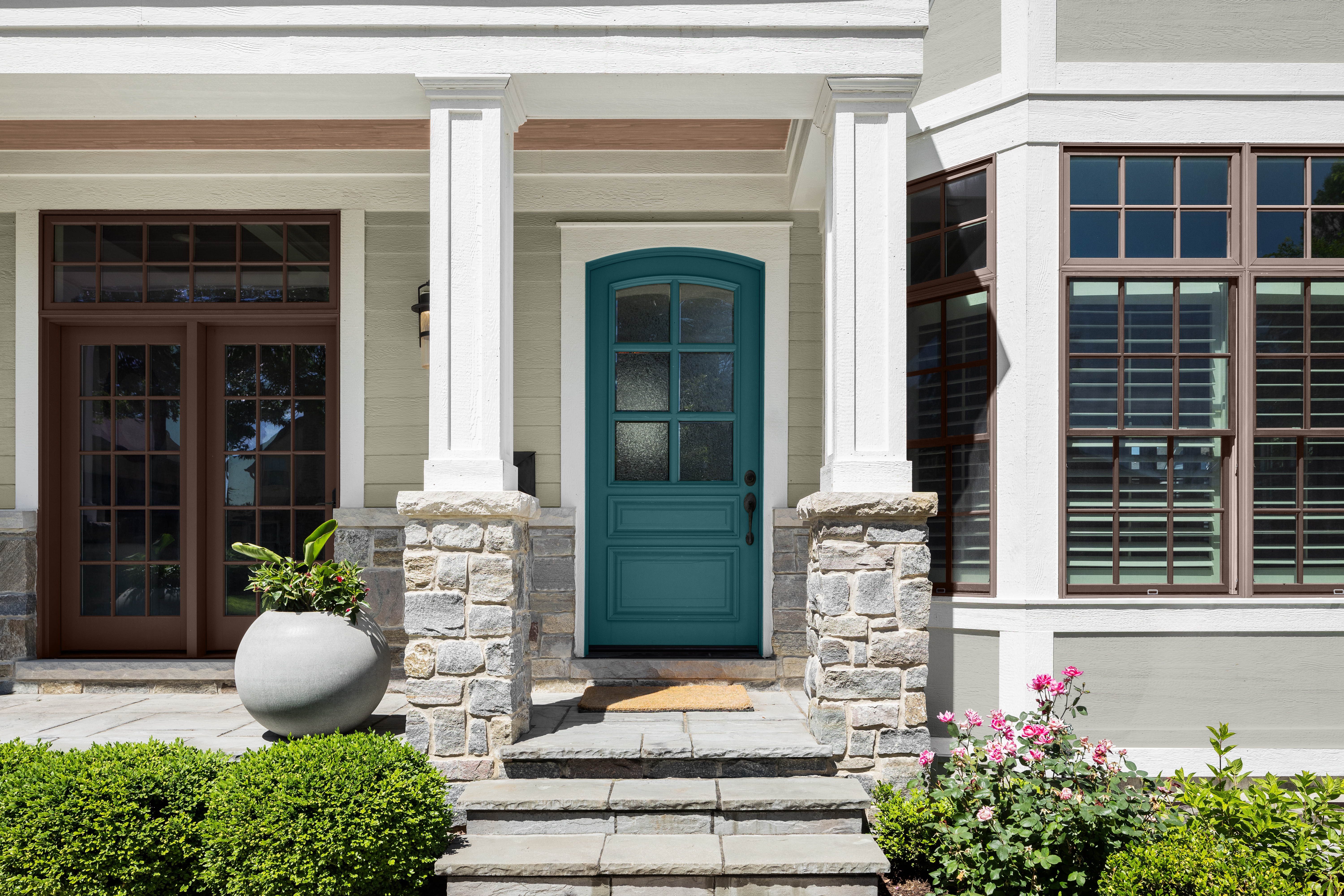 A front entry of a house with a deep sophisticated teal front door beneath a small, covered porch supported by white columns and stone bases. Stone steps lead up to the door, with a large grey planter on the left and landscaped shrubs and pink flowers in the foreground. Large windows with dark trim frame the entrance.