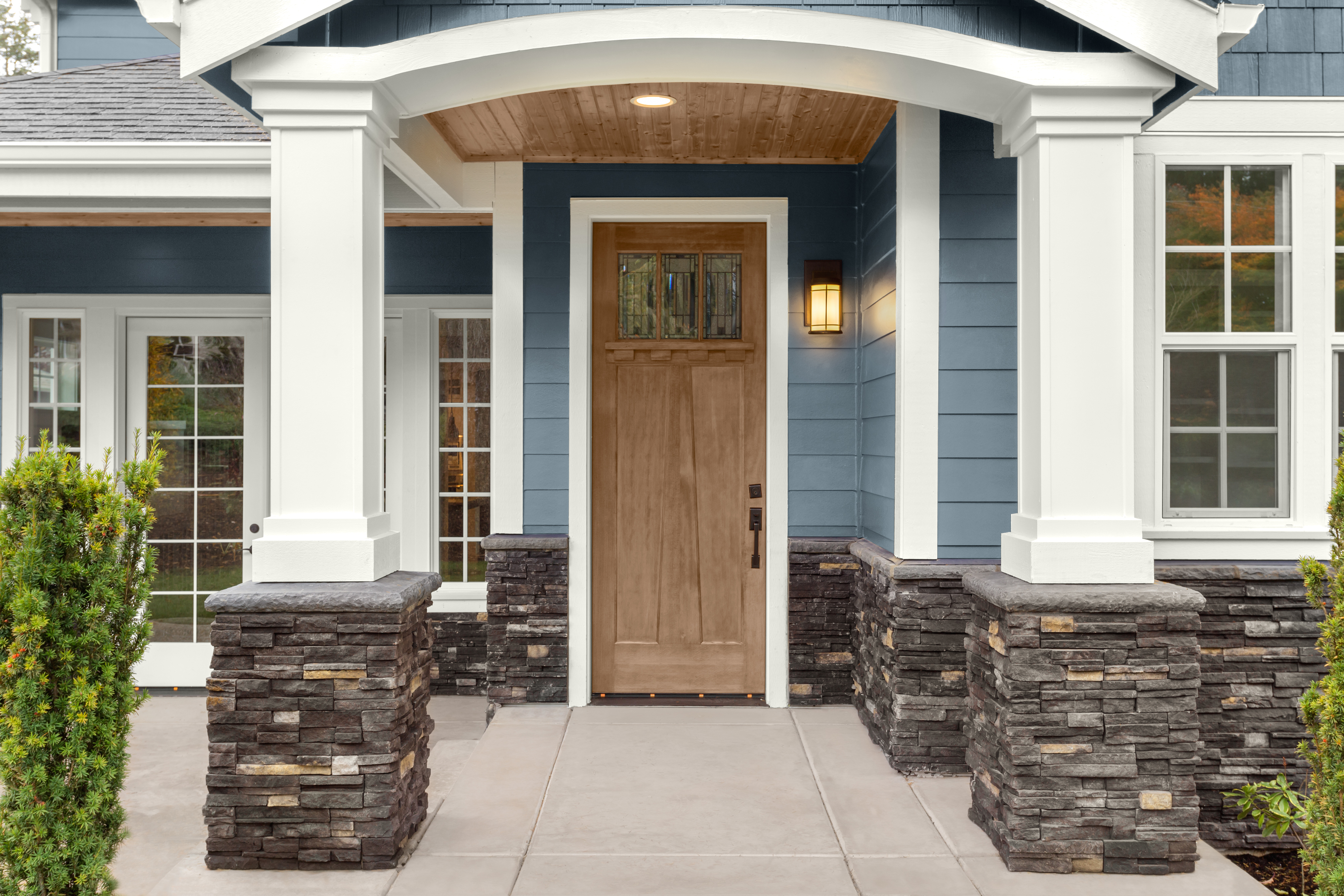 A front porch with a Taupe wood colour entry door, blue siding, and stacked stone columns, featuring a wood-stained ceiling under the covered entry. 