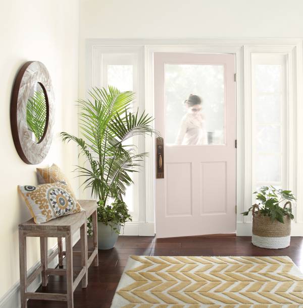 A bright entryway with white walls, a pale pink frosted-glass door with a faint figure outside, a small wooden bench with cushion and round mirror, potted plants, and a yellow chevron rug on dark wood floors.