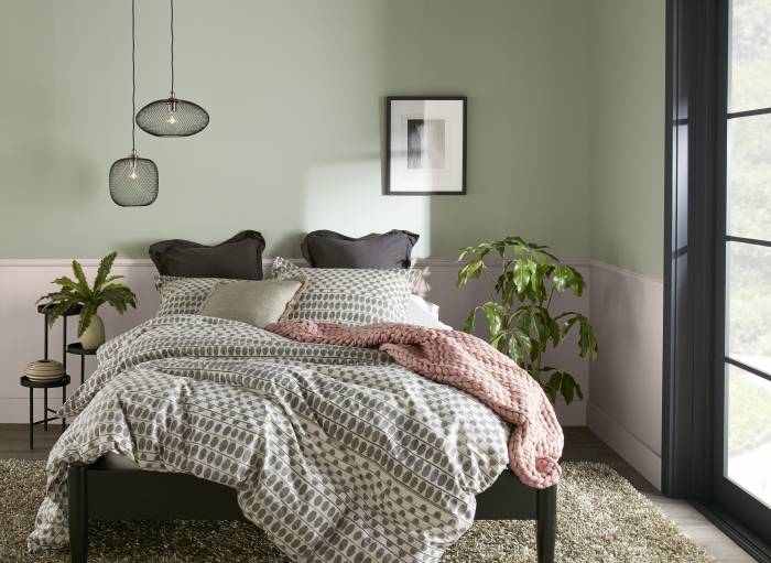 A serene bedroom with light pink and soft sage green walls and a dark bed frame dressed in patterned grey and white bedding. Layered pillows in neutral tones rest against the headboard, with a blush pink throw blanket draped across the bed. Two small pendant lights hang on one side, and a framed black-and-white artwork is centered above the bed.