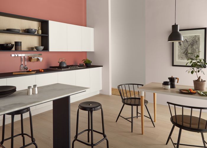 A modern kitchen and dining area with light pink walls and a terracotta accent wall above white upper cabinets and open wood shelving. A long grey countertop with black bar stools sits in the foreground, while a small light wood dining table with black wire chairs is placed nearby.