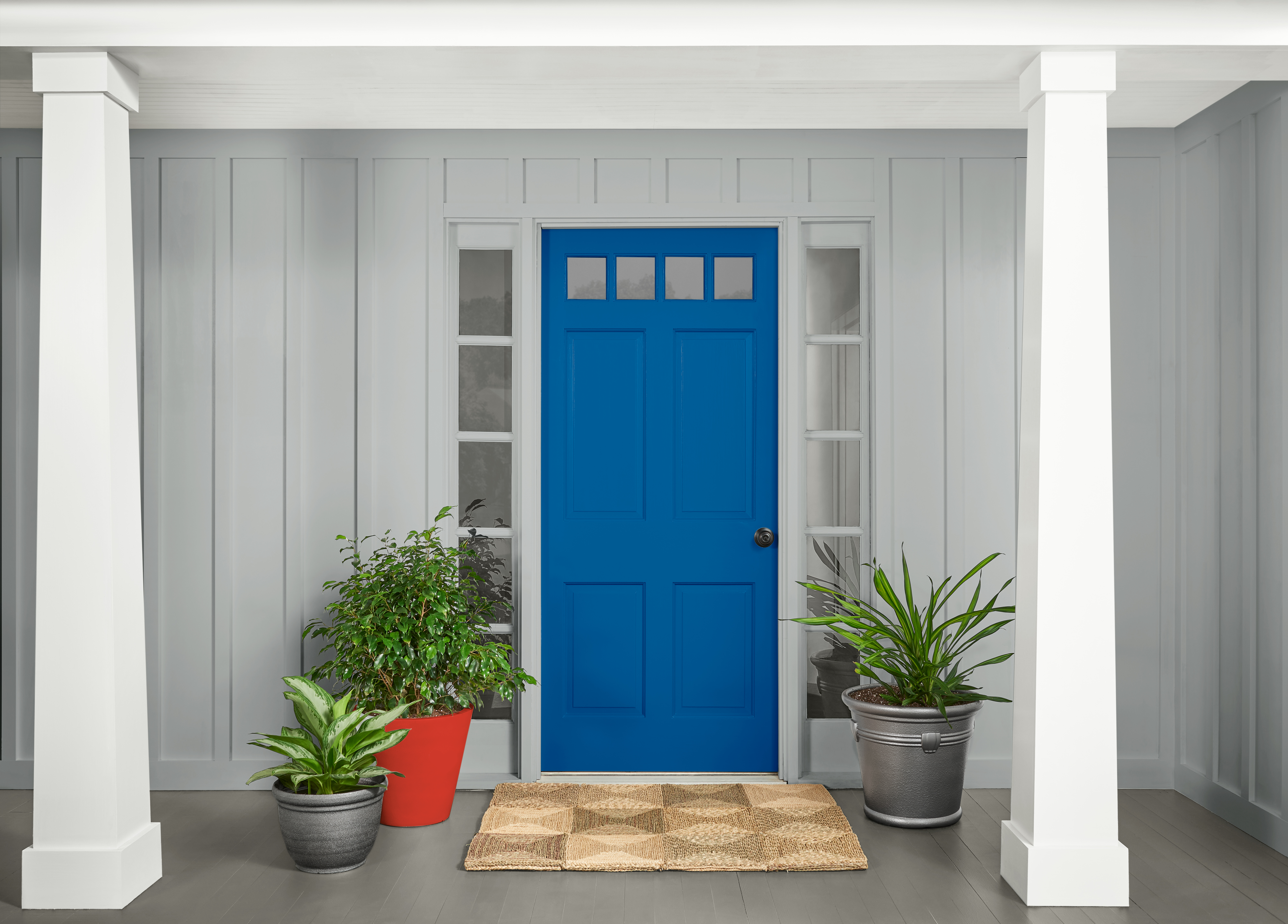 A front porch with light grey siding and white columns, featuring a bright blue front door centered between two narrow windows. Potted green plants sit on either side of the door, and a woven doormat rests on the grey floorboards.