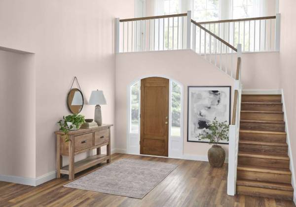 An airy high ceiling foyer with light pink walls, a wooden front door framed by arched windows, a staircase with walnut and white railing, a console table with lamp and plants, and a neutral area rug on wood floors.